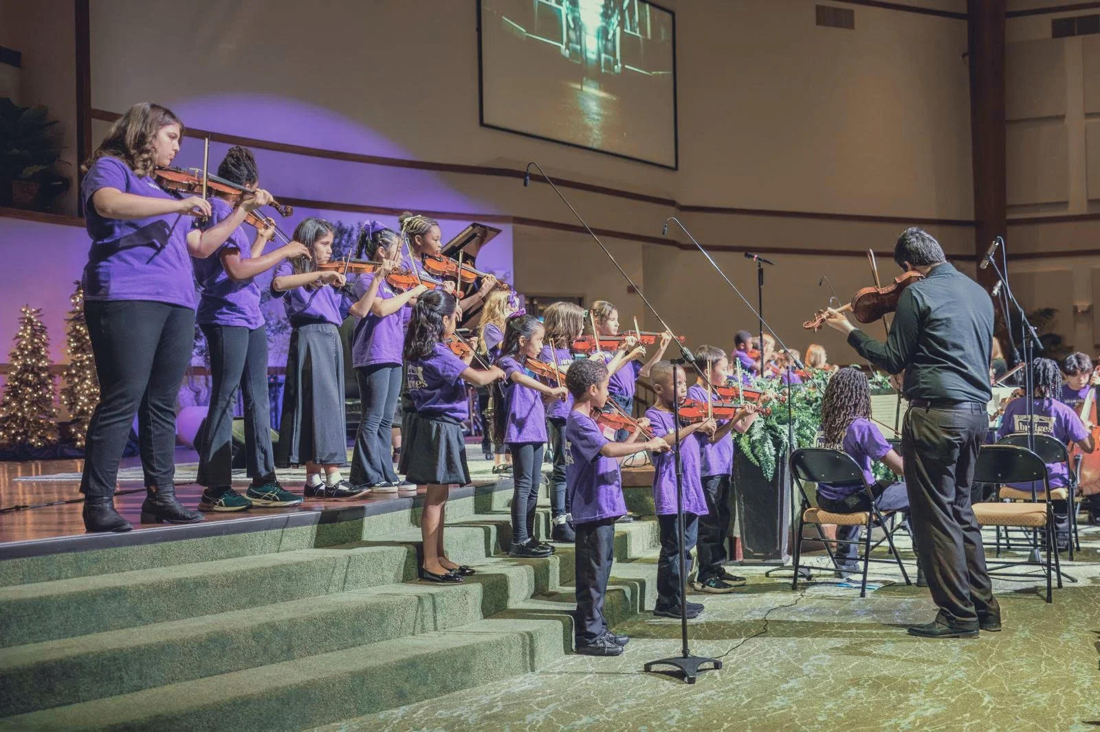 Children and adults playing violins during a church choir concert, with Christmas decorations and a Christmas tree in the background.