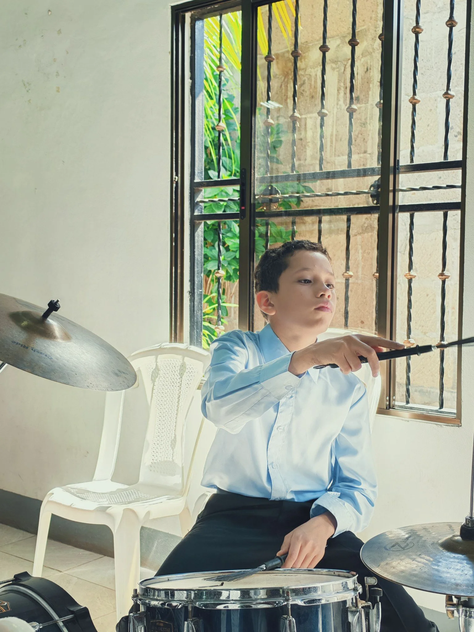 Young boy with short dark hair wearing a light blue dress shirt playing a drum set indoors near a window with black metal bars. White plastic chair and a white basket are in the background. Green plants visible outside the window.