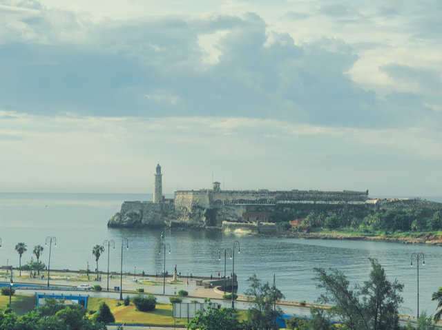 A lighthouse and historic fortress on a rocky coastline, with a park and walking paths in the foreground, and cloudy skies above.