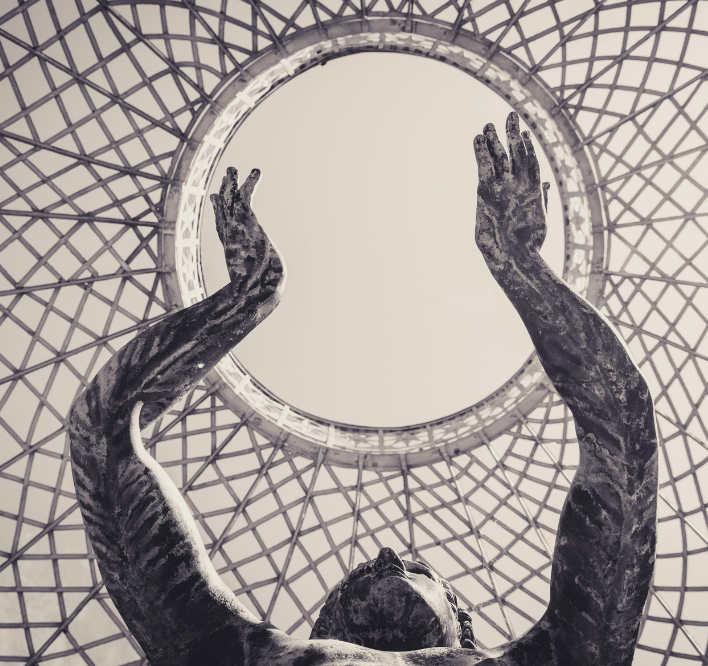 A statue of a person reaching upward with both arms, viewed from below against a modern, intricate geometric ceiling structure with a circular opening.