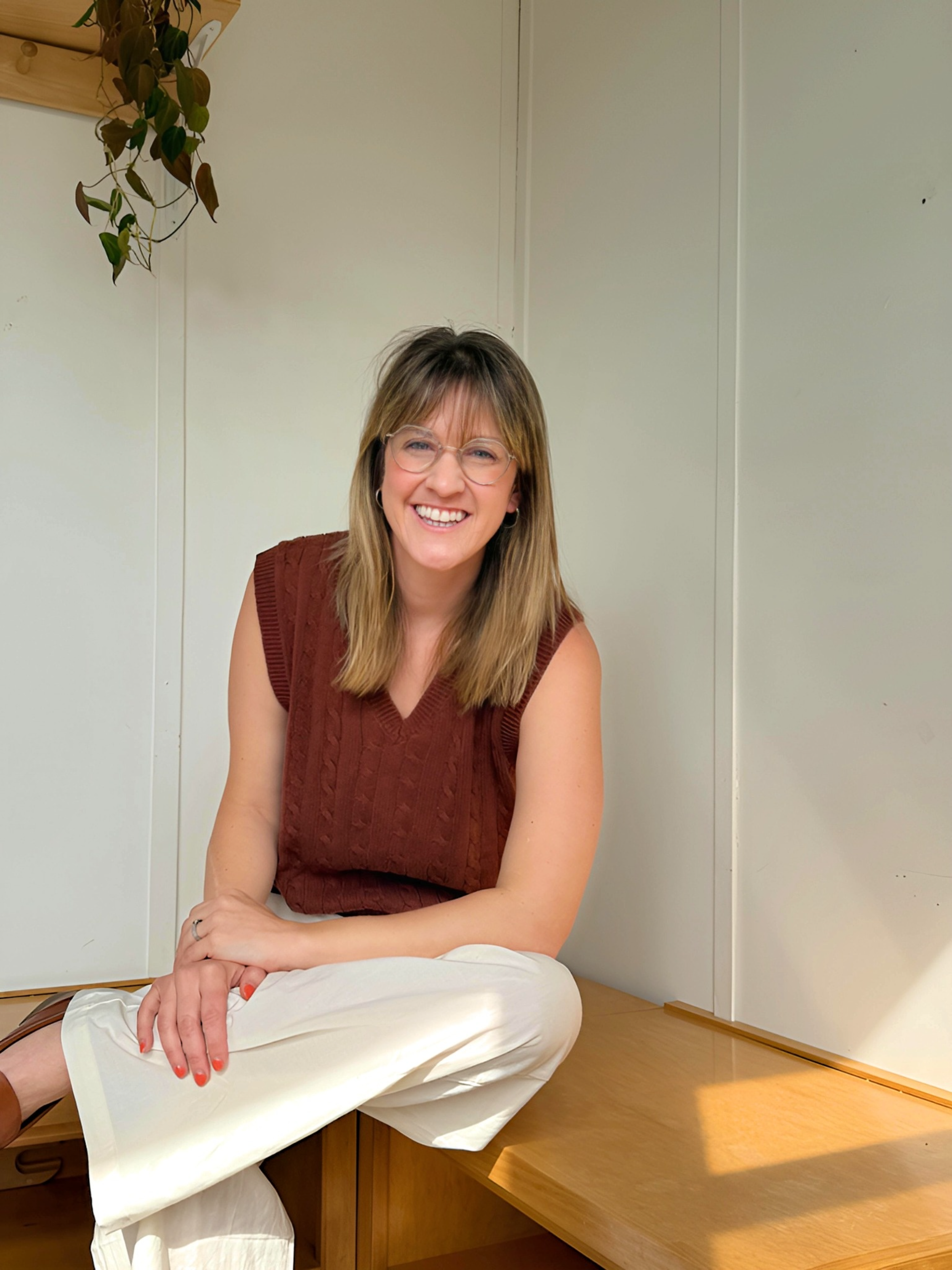 A woman with shoulder-length light brown hair, wearing glasses, a brown sleeveless top, and cream-colored pants, sitting on a wooden bench, smiling at the camera in a room with white walls.