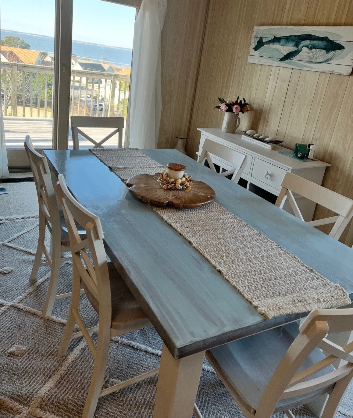 A dining room with a large wooden table featuring a woven table runner and a decorative candle centerpiece. Four white chairs are positioned around the table. In the background, a sliding glass door opens to a balcony with a view of water and rooftops. A white sideboard and a wall art of a whale are also visible, with a vase of flowers on the sideboard.
