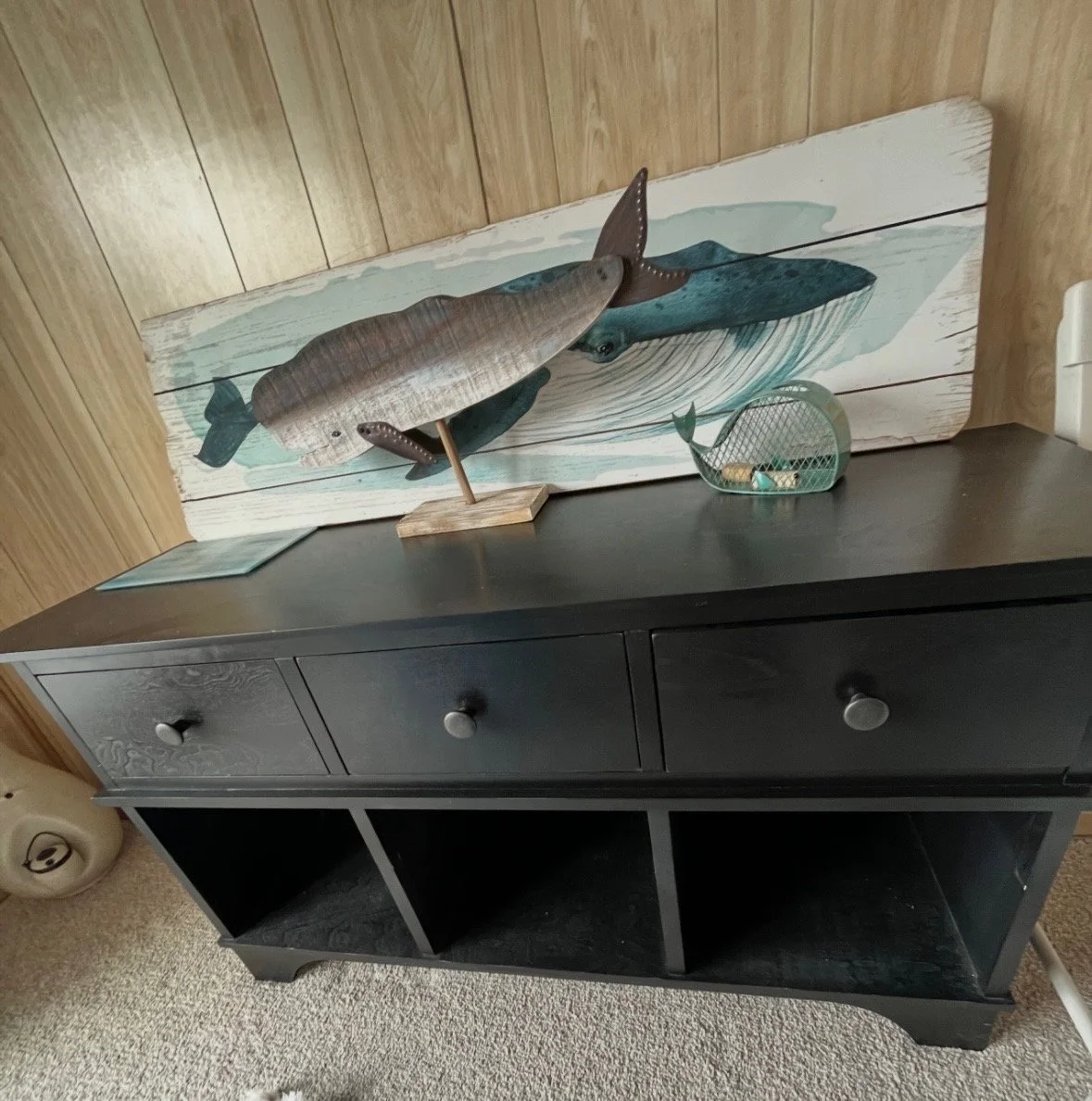 Decorative art piece on a black cabinet, featuring a large painted wooden whale with smaller wooden whale figures and a slender wooden whale sculpture on a stand in front.