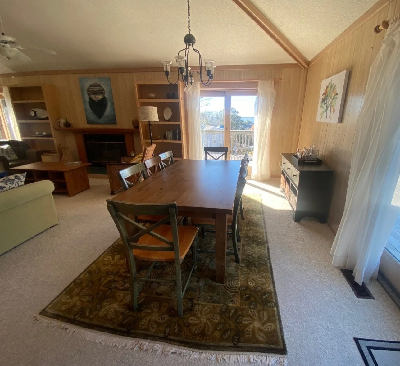 Dining room featuring a wooden table with six chairs, a rug underneath, and a sideboard along the wall. Sunlight streams through sliding glass doors with sheer curtains, leading to an outdoor deck. There is a living area with a fireplace, built-in shelves, and a ceiling fan.