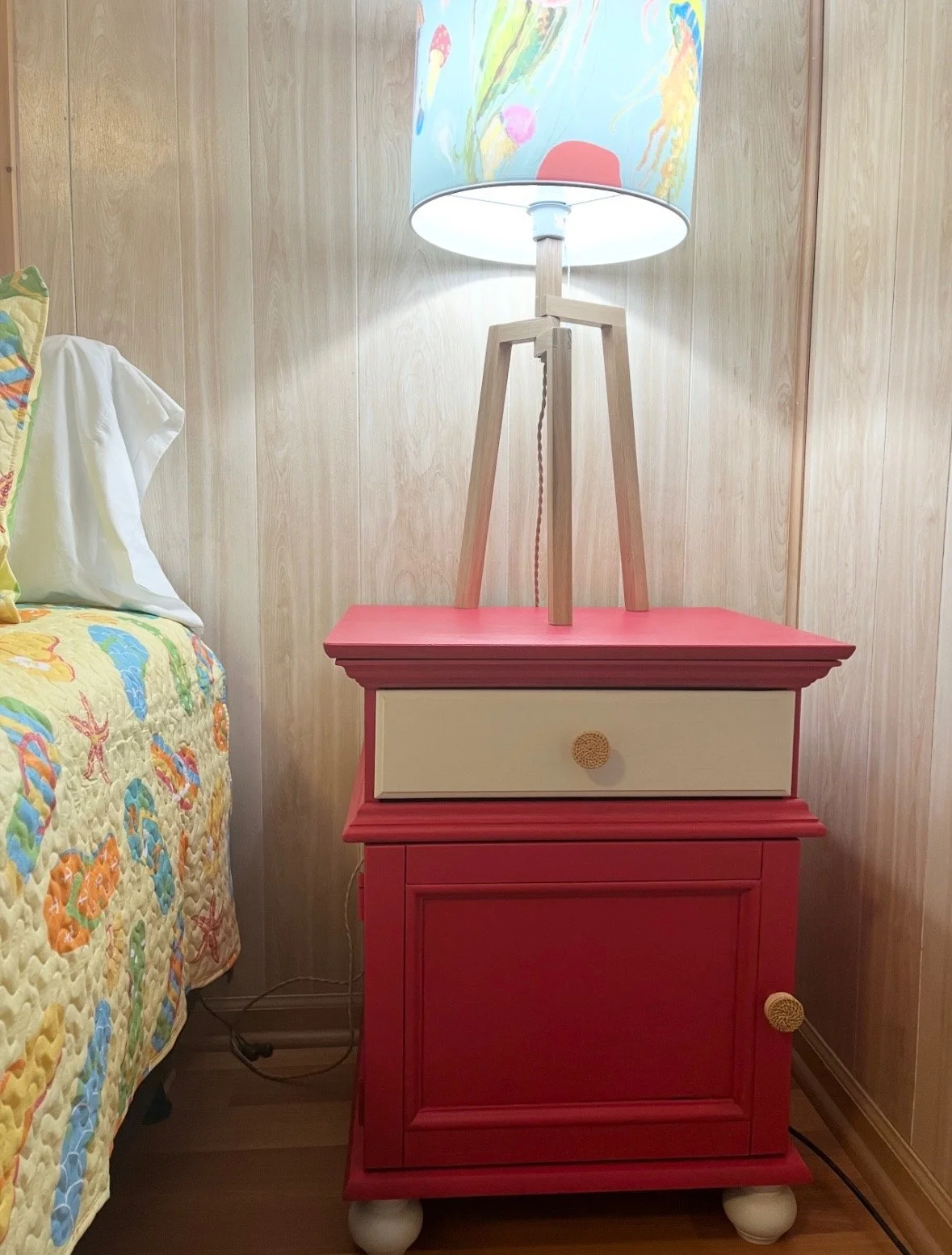 A pink and cream-colored wooden nightstand with a colorful lamp on top, placed next to a bed with a sea-themed quilt and pillows in a room with wooden panel walls.