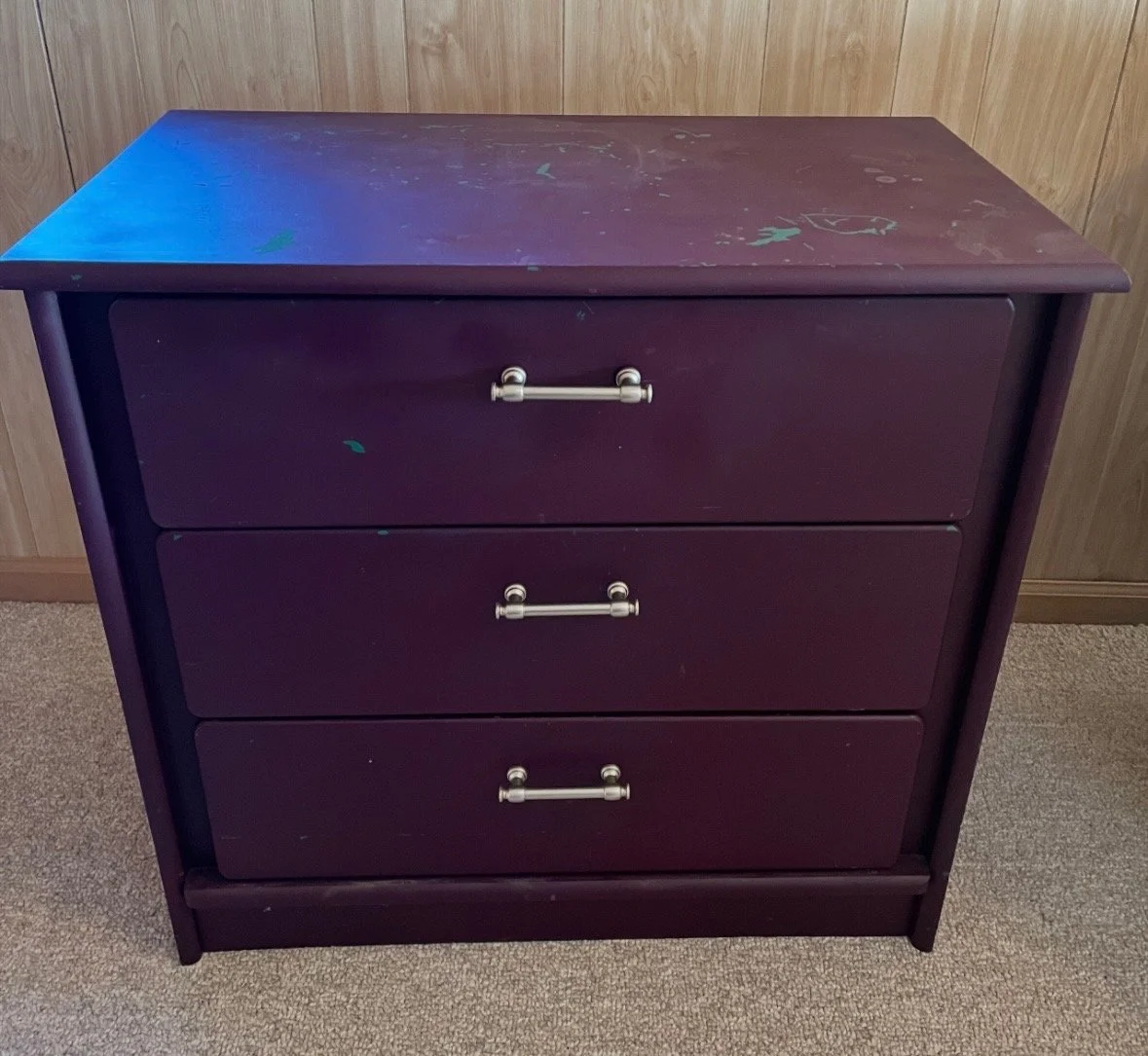 A purple wooden dresser with three drawers, silver handles, and scratches on the top surface, located against a wood-paneled wall on a beige carpet.