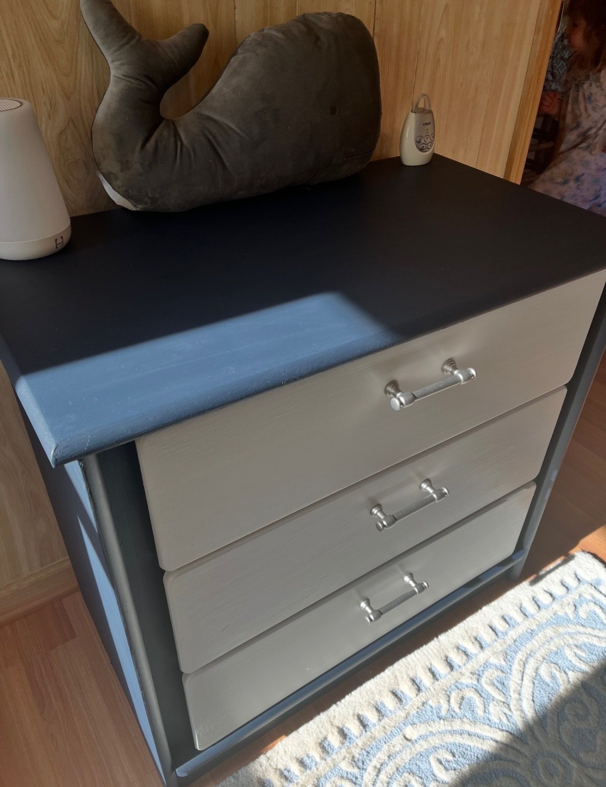 A bedside table with a black top, three beige drawers with silver handles, a whale-shaped pillow, two bedside lamps, a wooden wall, and a partially visible person in patterned clothing.