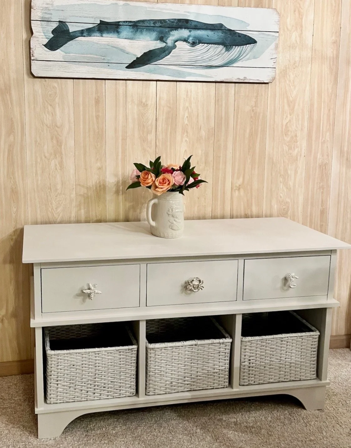 Light-colored dresser with three drawers and three woven baskets underneath, decorated with a ceramic vase of pink and orange roses, a whale painting above, against a wooden wall and carpeted floor.