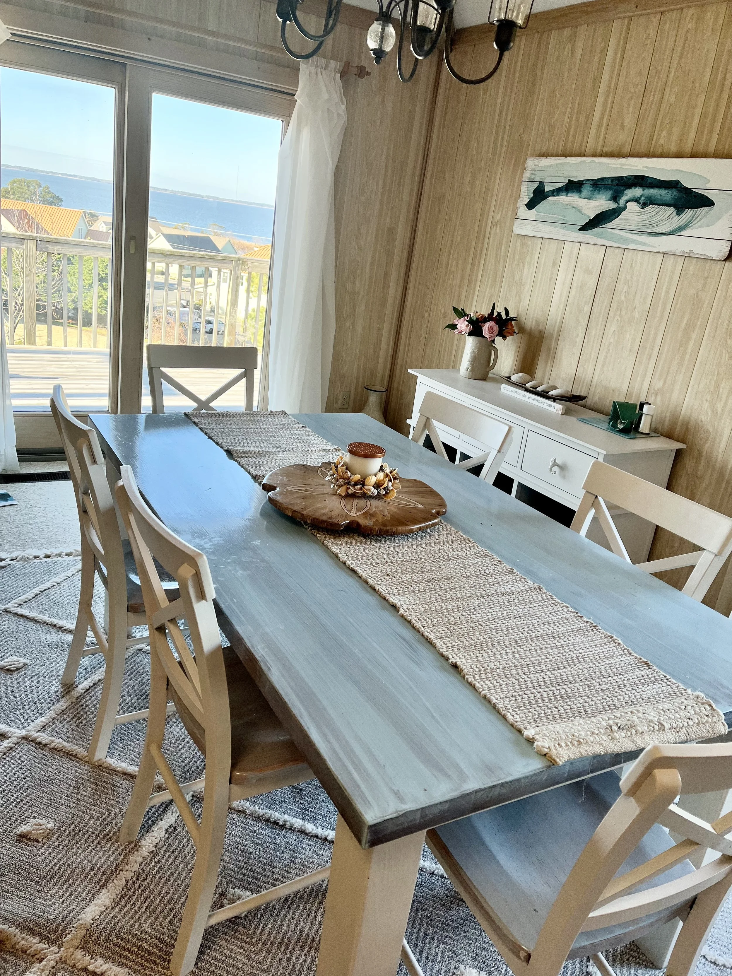Dining room with a large wooden table, six chairs, a buffet with a vase of pink flowers, a wall art of a whale, and a view of a balcony overlooking water through glass sliding doors.