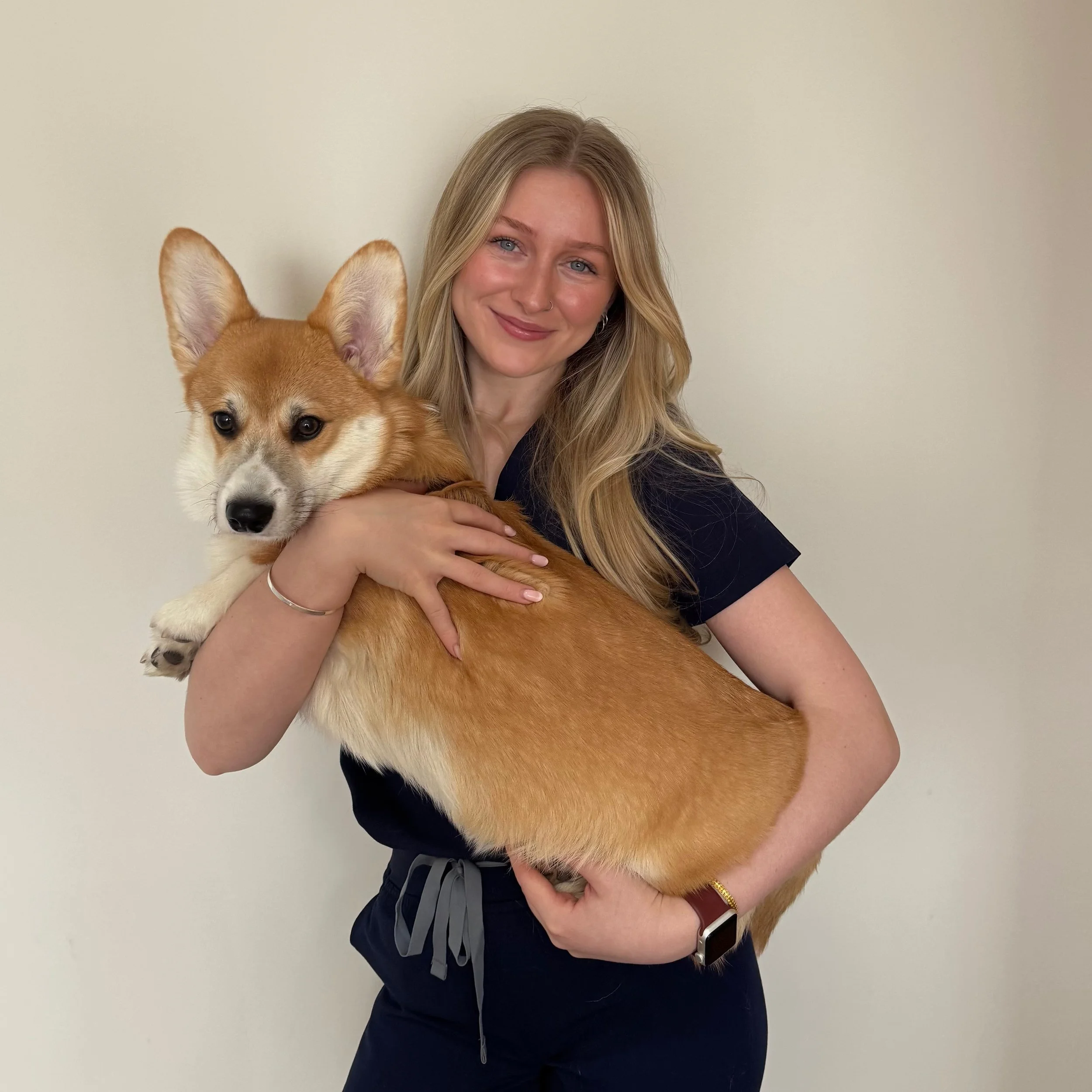 A young woman with long blonde hair, blue eyes, and a nose ring hugging a corgi dog with a fluffy orange and white coat against a plain off-white wall.
