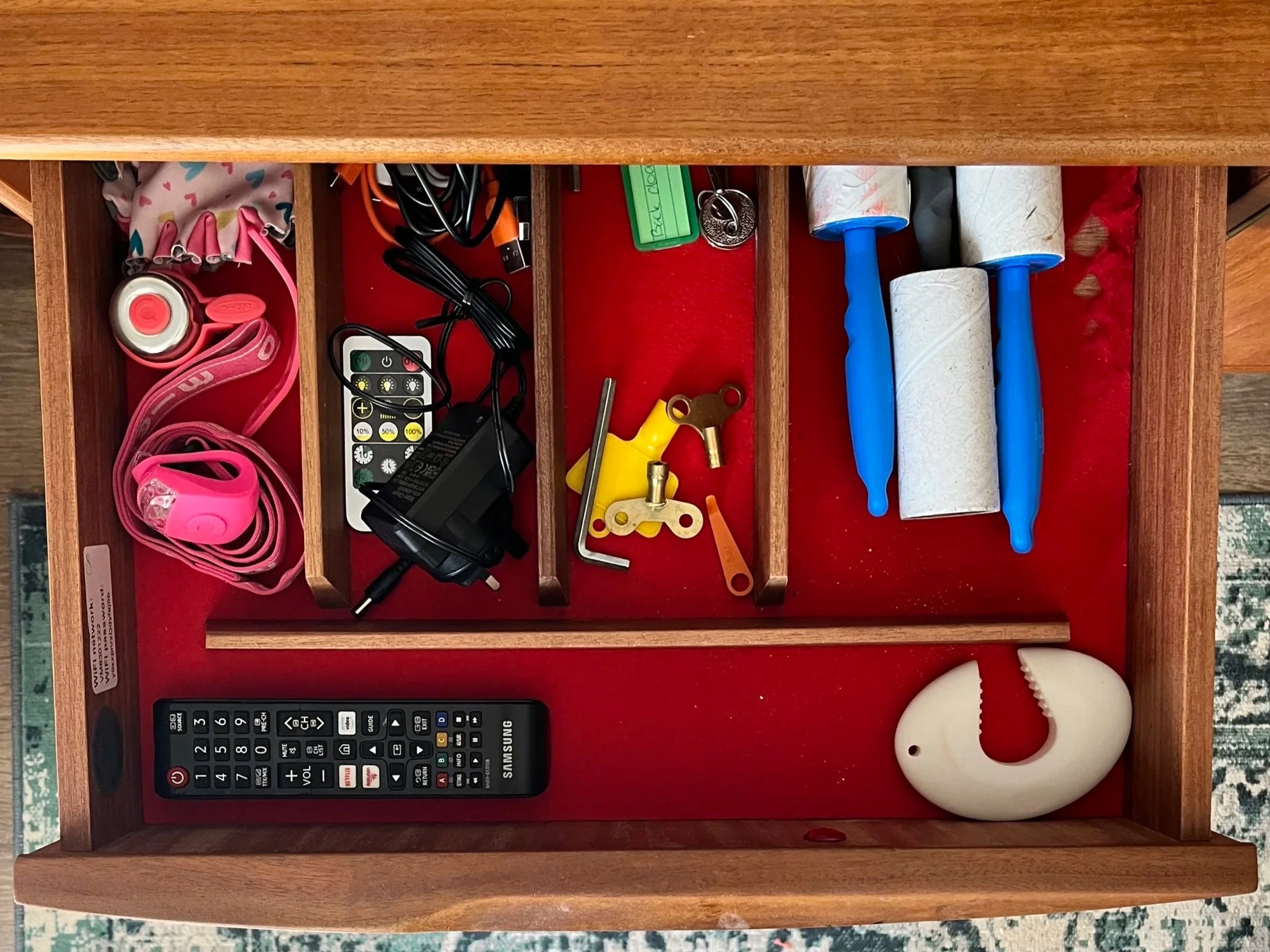 A wooden drawer with red lining containing various office and household items such as a pink headband, earphones, a remote control, a small yellow toy, rulers, keys, duct tape rolls, spray bottles, scissors, and a plastic shoehorn.