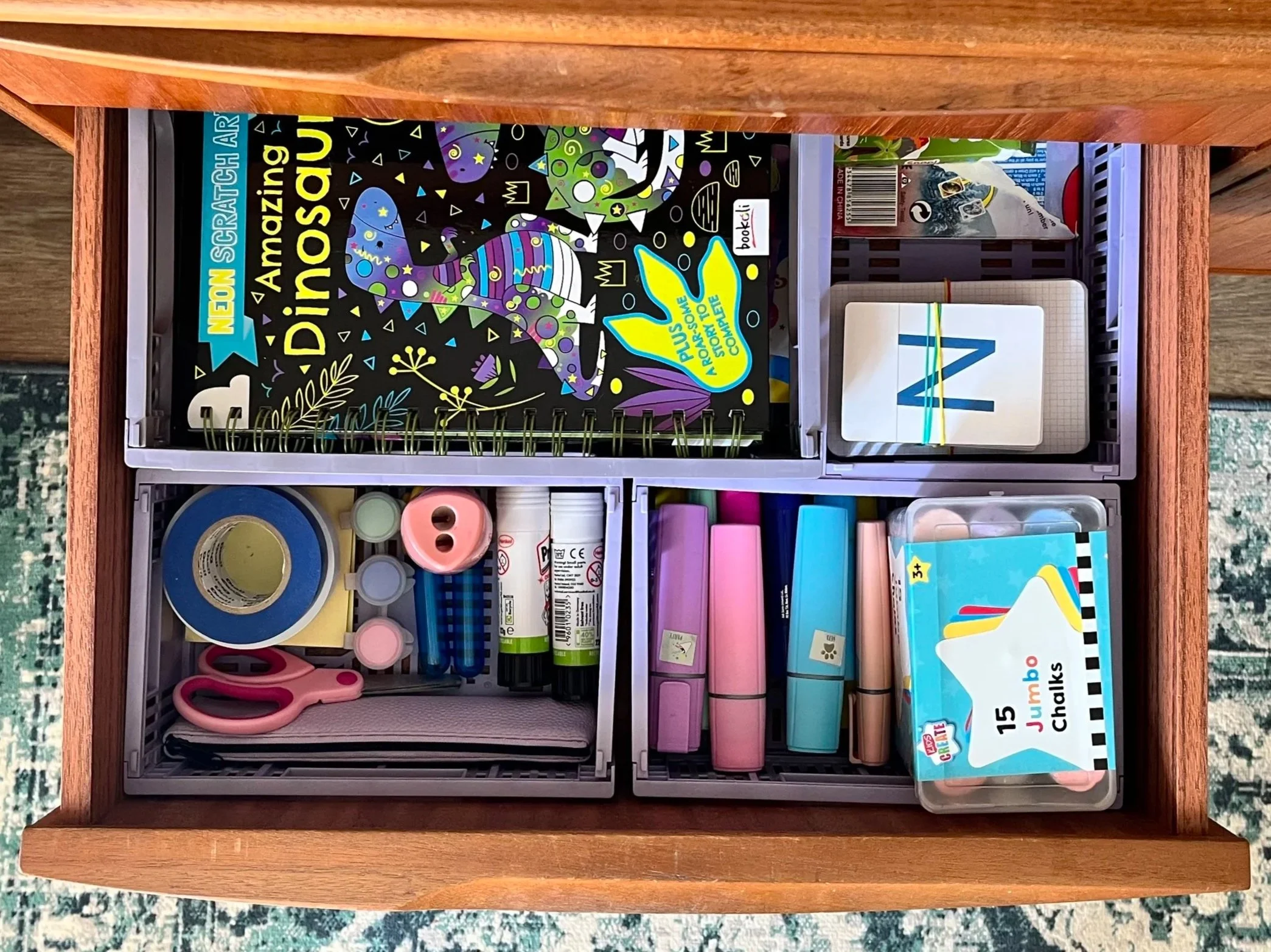 A wooden drawer filled with school supplies, including a colorful book titled 'Amazing Dinosaur' with a cartoon dinosaur illustration on the cover, rolls of tape, pink scissors, glue sticks, and a pack of jumbo chalks.