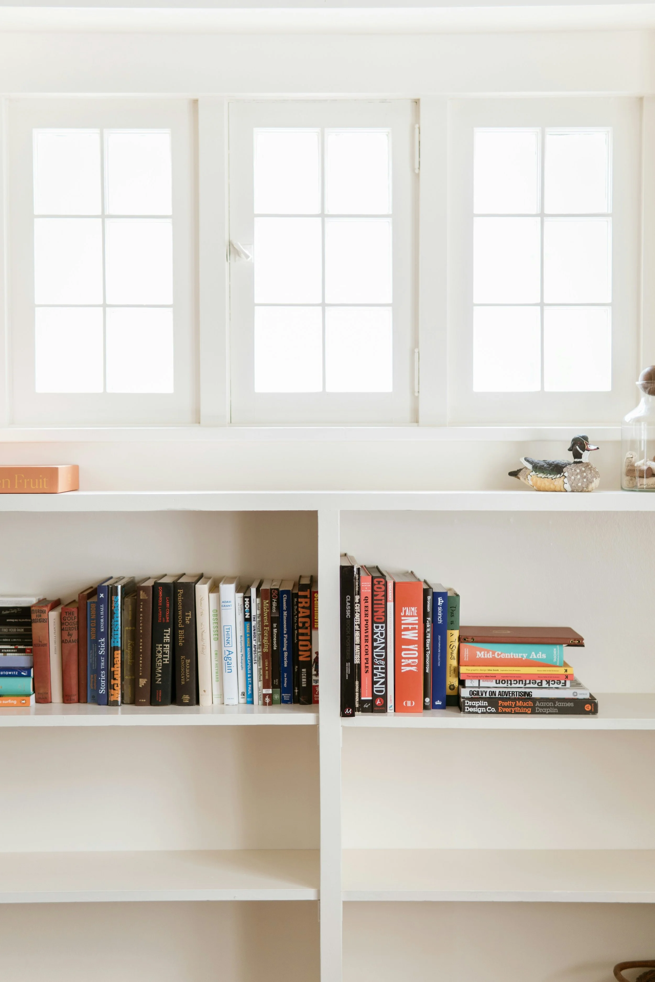 White built-in bookshelf with two shelves filled with a variety of books, a duck figurine, glass jars, and a small stack of colorful books on the right.