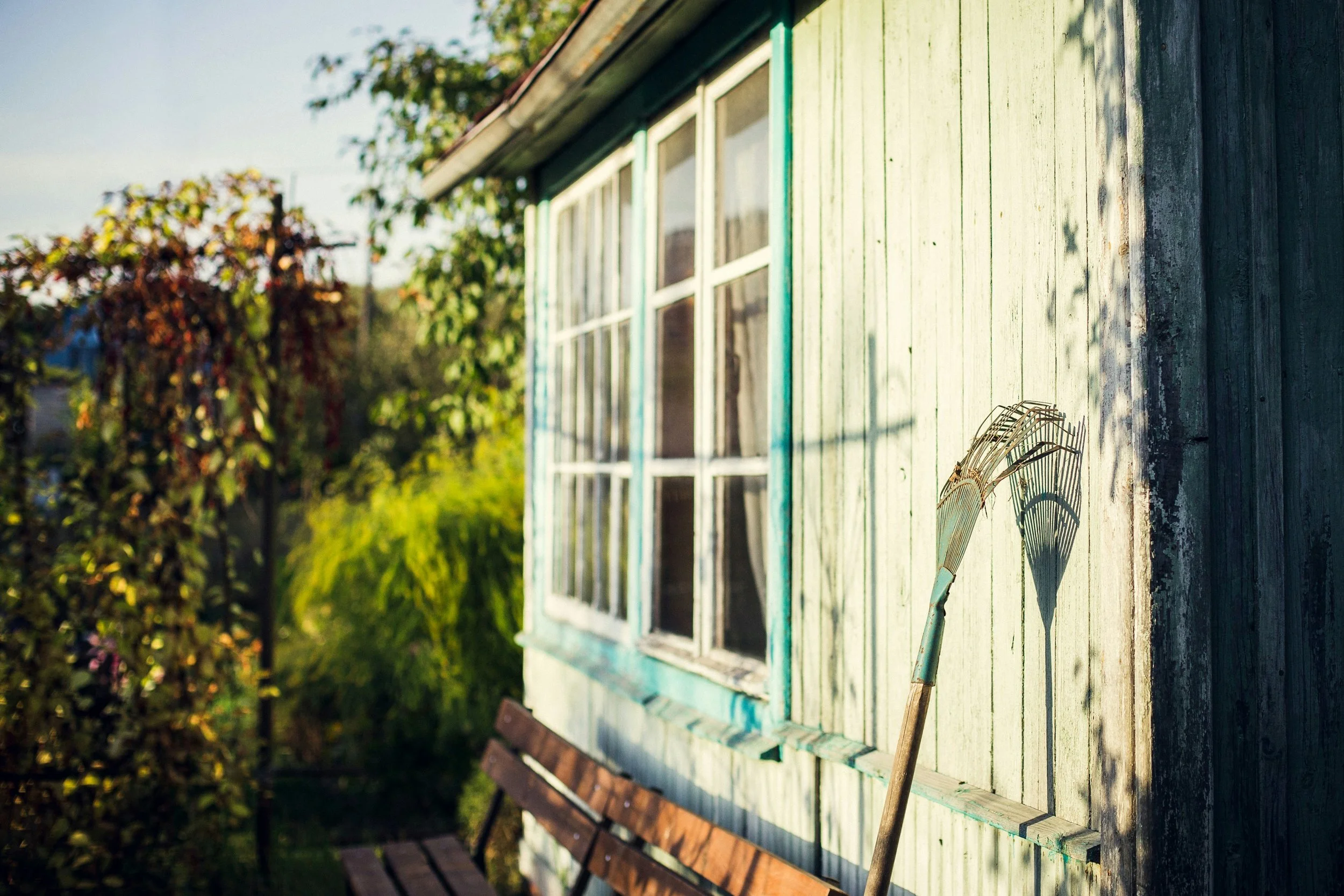 A weathered, green-painted house with white window frames viewed from the side in a garden with trees and a seating area. A gardening rake leans against the house.