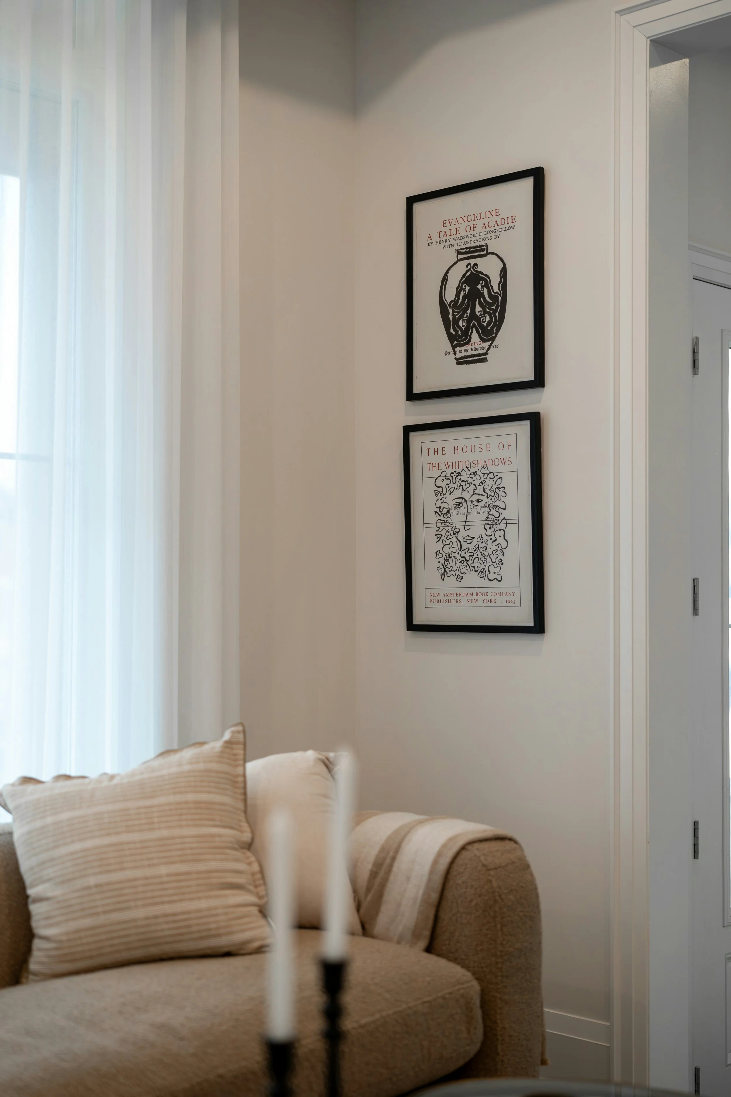 Living room corner with beige couch and striped pillows, two framed art prints on the wall, sheer white curtains, and a collection of candles in the foreground.