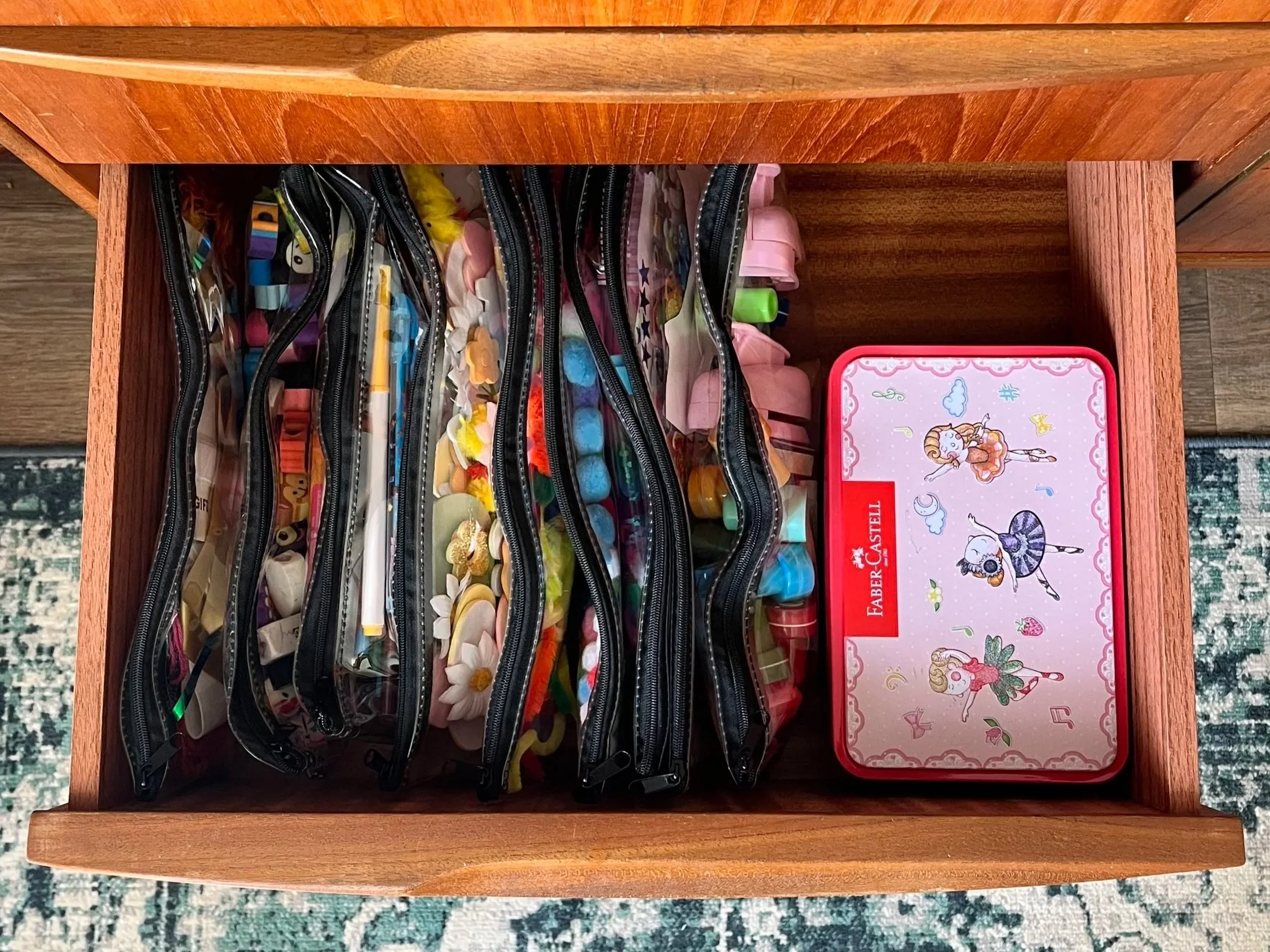 Open wooden drawer containing multiple clear plastic zipper pouches filled with various small items, and a pink box from the brand Faber-Castell with unicorn illustrations.