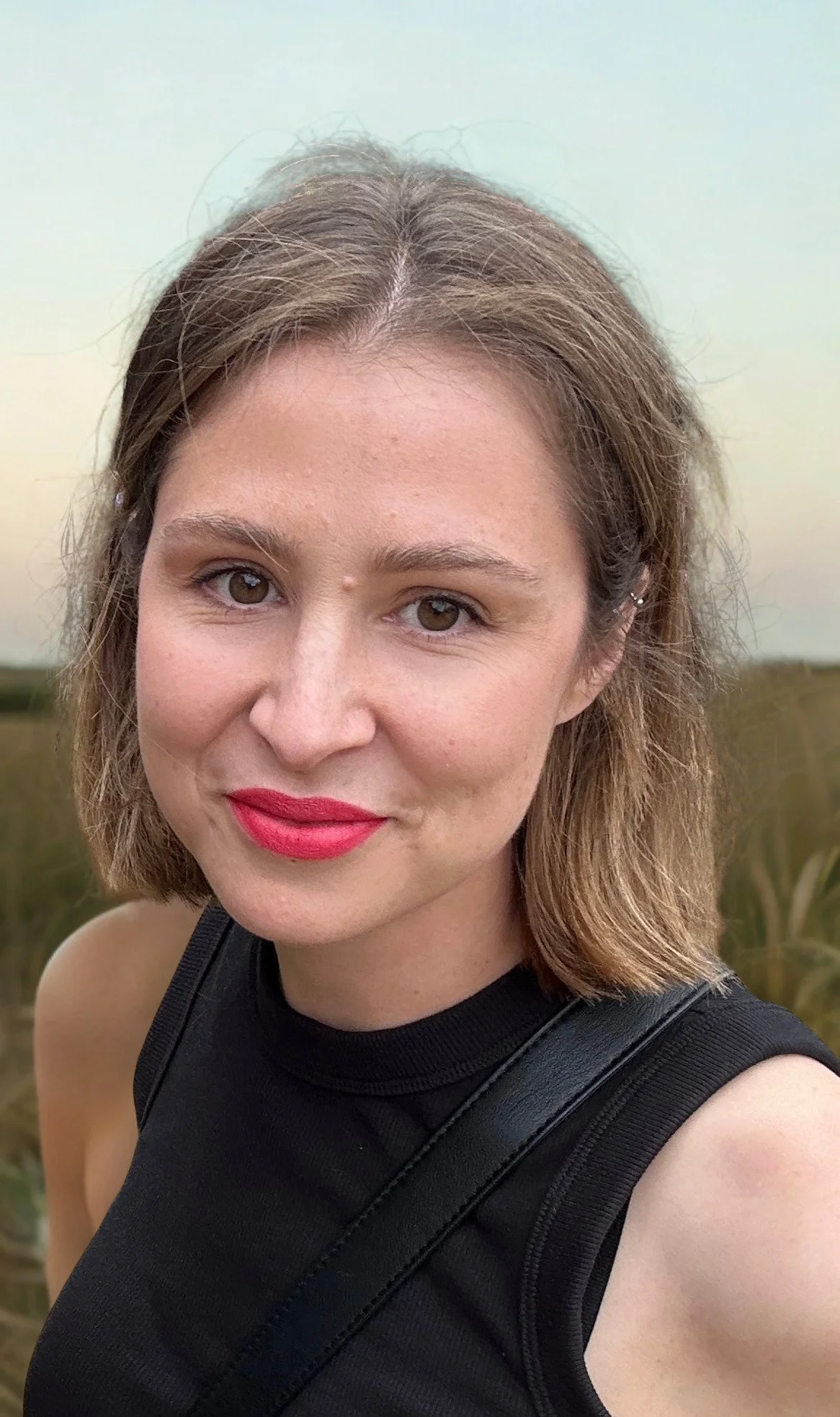 A woman with shoulder-length brown hair, light skin, red lipstick, and a black sleeveless top standing outdoors in a natural setting.