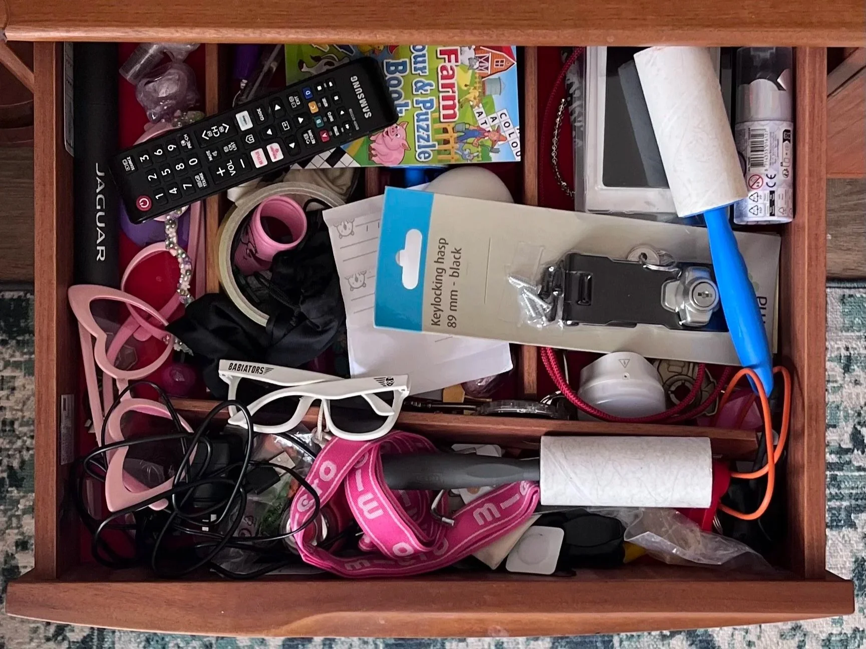 This is an open drawer filled with miscellaneous items, including a TV remote, children's glasses, a key lock, tape, a roll of paper towels, a book titled 'Peppa Pig: Fun and Learning,' various cords and electronic accessories, a headband, a small white box, and other various objects.