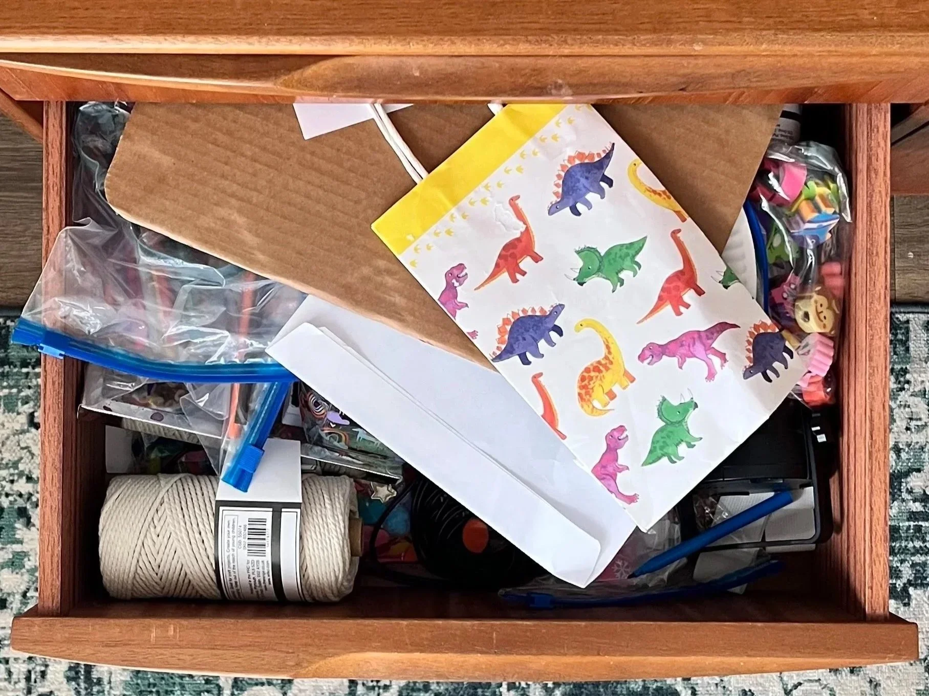 An open drawer containing various craft supplies, including a sheet of paper with colorful dinosaur stickers, a roll of beige twine, plastic bags with small items, and writing utensils.