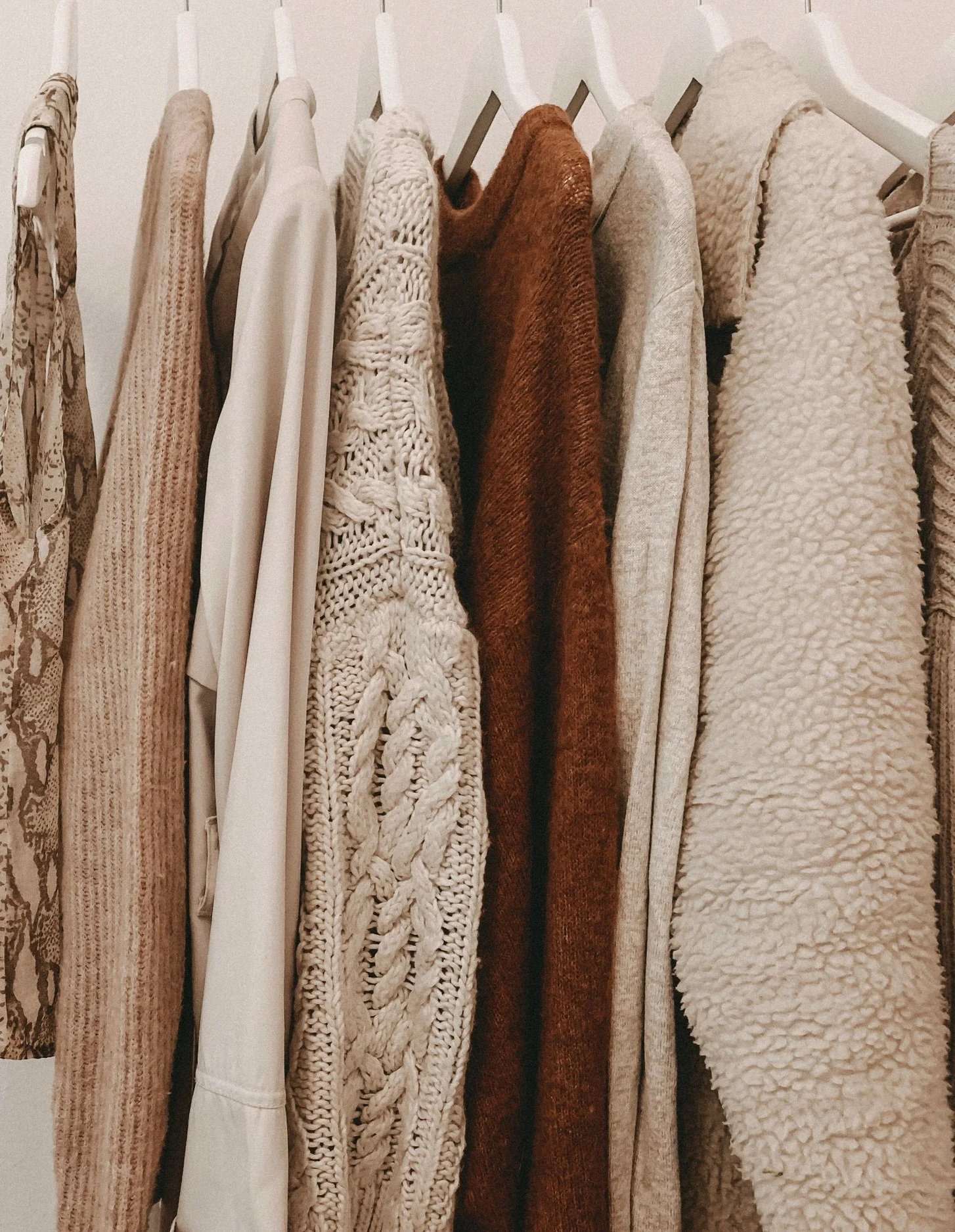 A row of various beige, white, and brown sweaters and cardigans hanging on white hangers on a clothing rack.