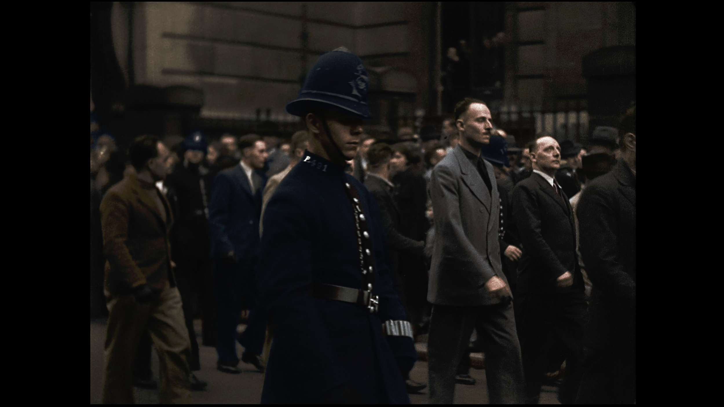 A crowd of people walking in an urban setting at night, including a police officer in uniform and a man in a gray suit.