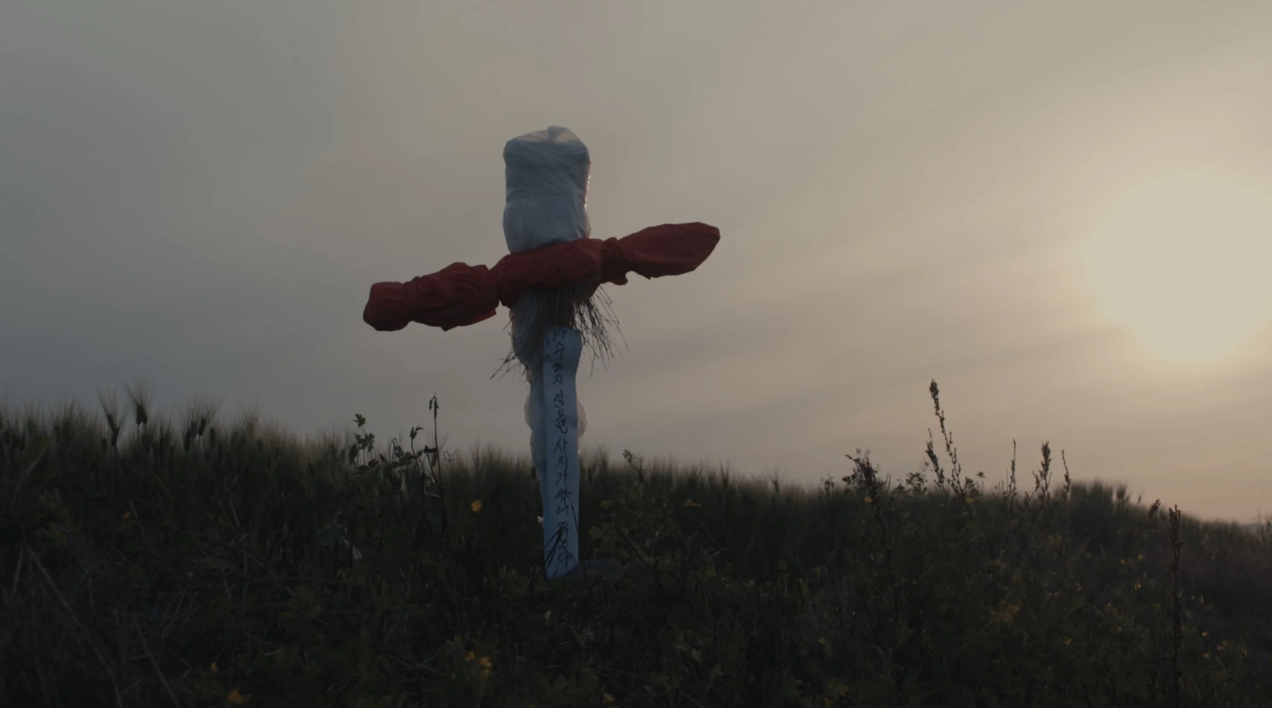 A roadside cross with a cloth tied around it and a cloth covering the top, set against a sunset sky with grassy terrain in the foreground.