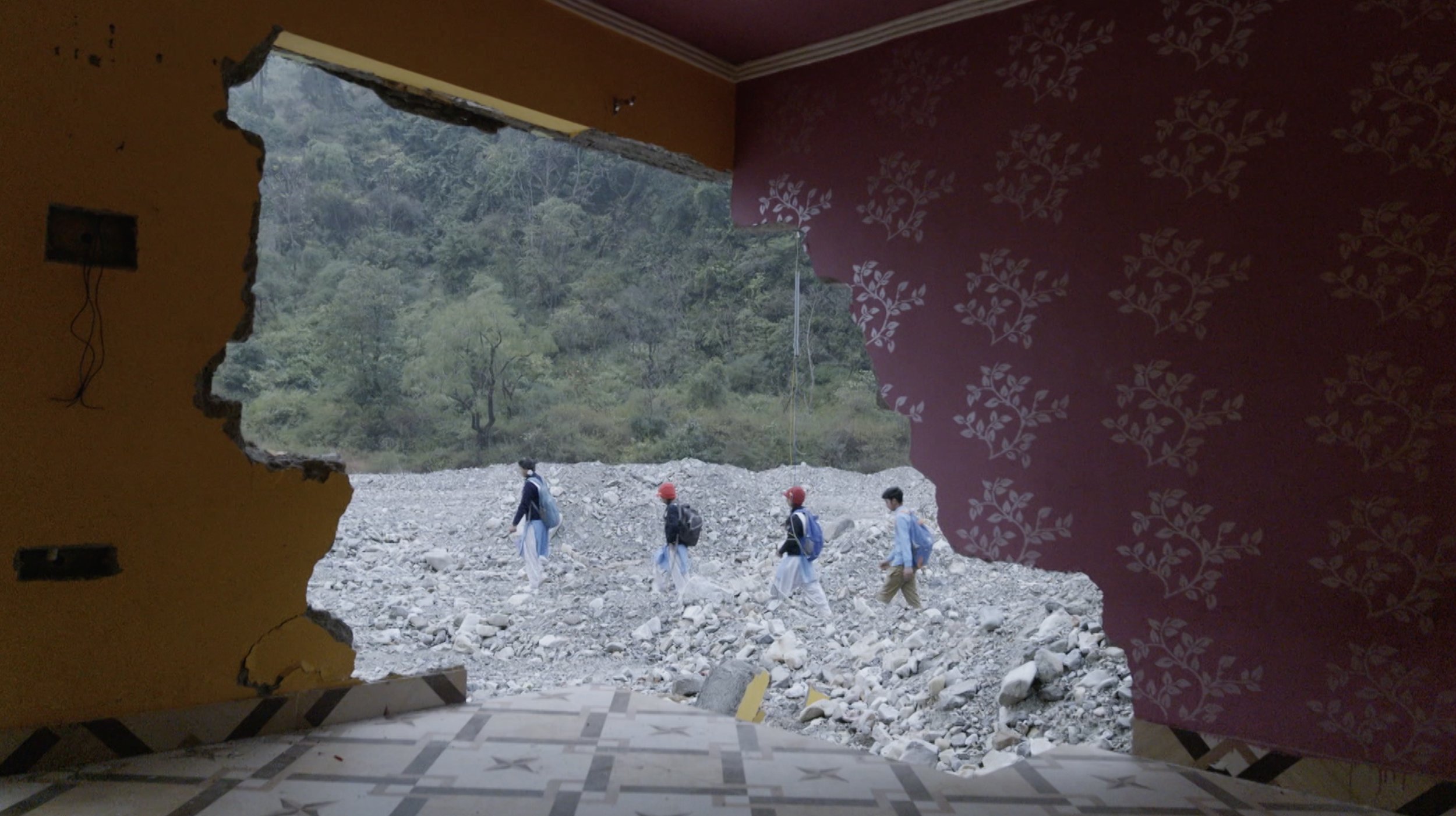 View through a large hole in the wall showing a rocky landscape and a group of people walking outdoors, with mountains and trees in the background.