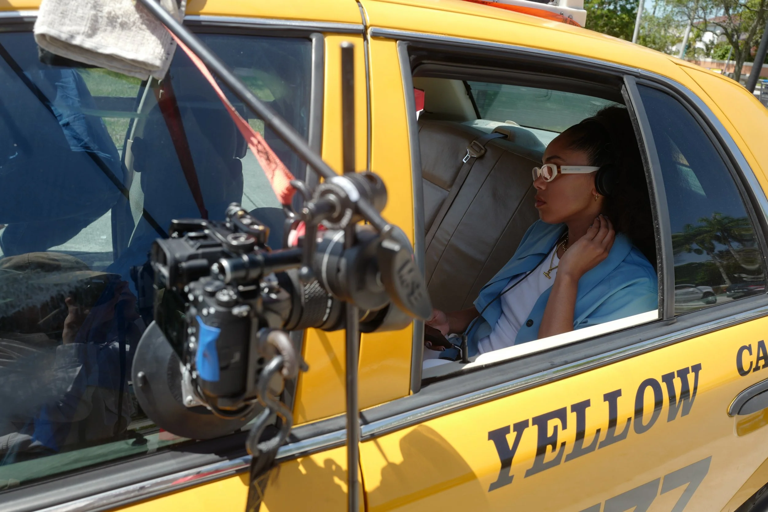 A woman inside a yellow vehicle, possibly a van, wearing glasses and a blue and white uniform, sits in the driver's seat with her hand near her ear, as a camera or filming equipment is positioned in front of her.