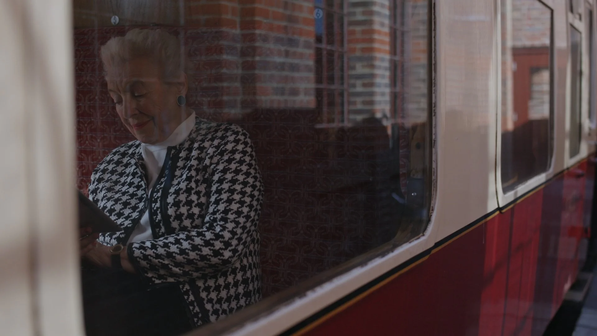 An elderly woman with short, blonde hair, wearing a patterned black and white jacket and white blouse, sits inside a vintage train, visible through a window with a brick building reflected behind her.