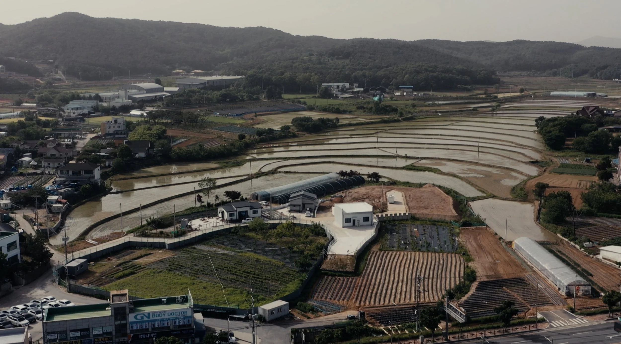 Aerial view of terraced rice fields, small houses, greenhouses, and surrounding mountains in a rural area.
