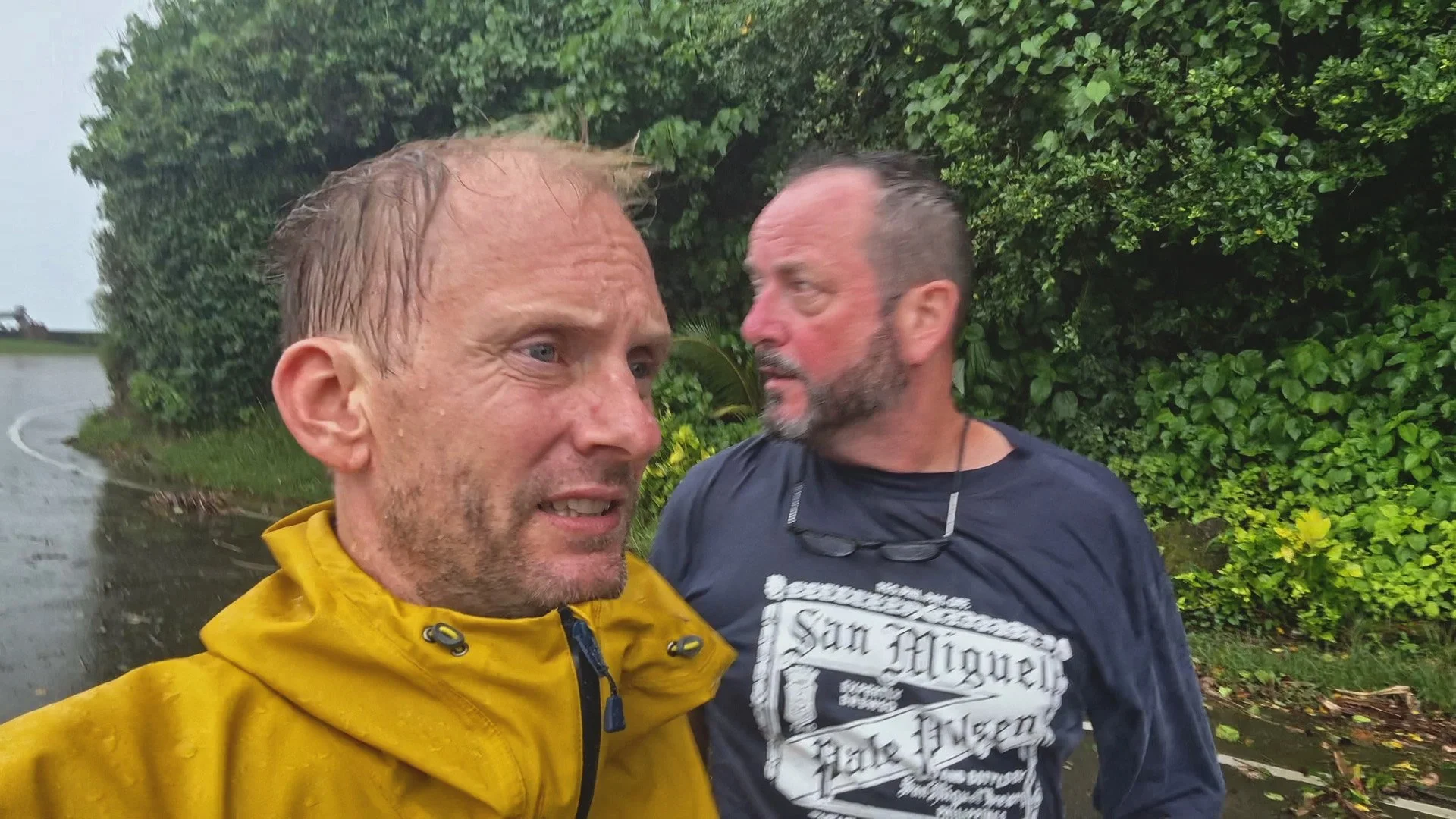Two men are outdoors on a rainy day, standing close to each other. The man in the foreground is wearing a yellow rain jacket with water droplets on it, and the man in the background is wearing a black T-shirt with white text. They are near green bushes, and rain is visible on the ground and in the air.