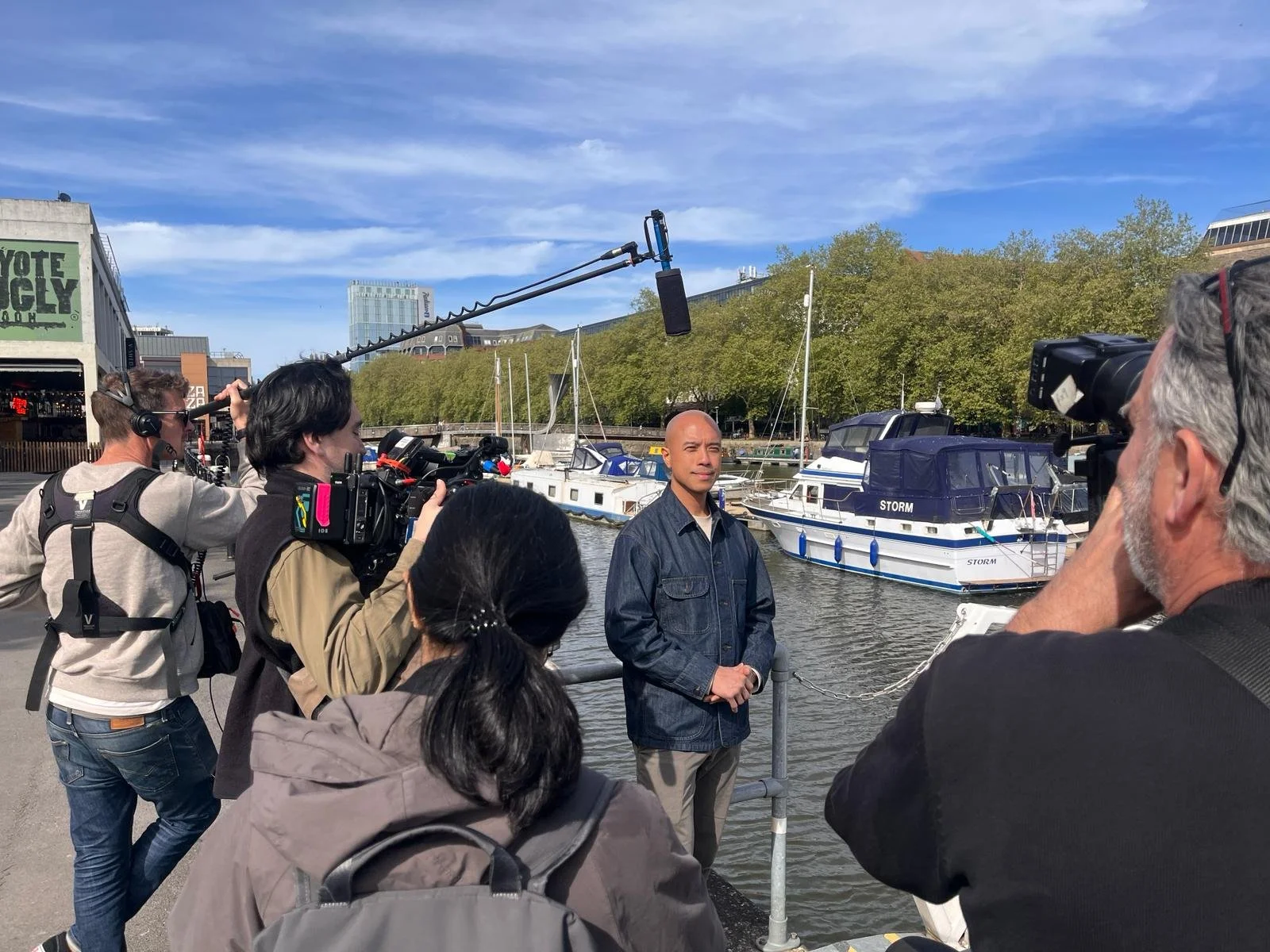 Group of people filming a man by the water near boats and trees under a blue sky