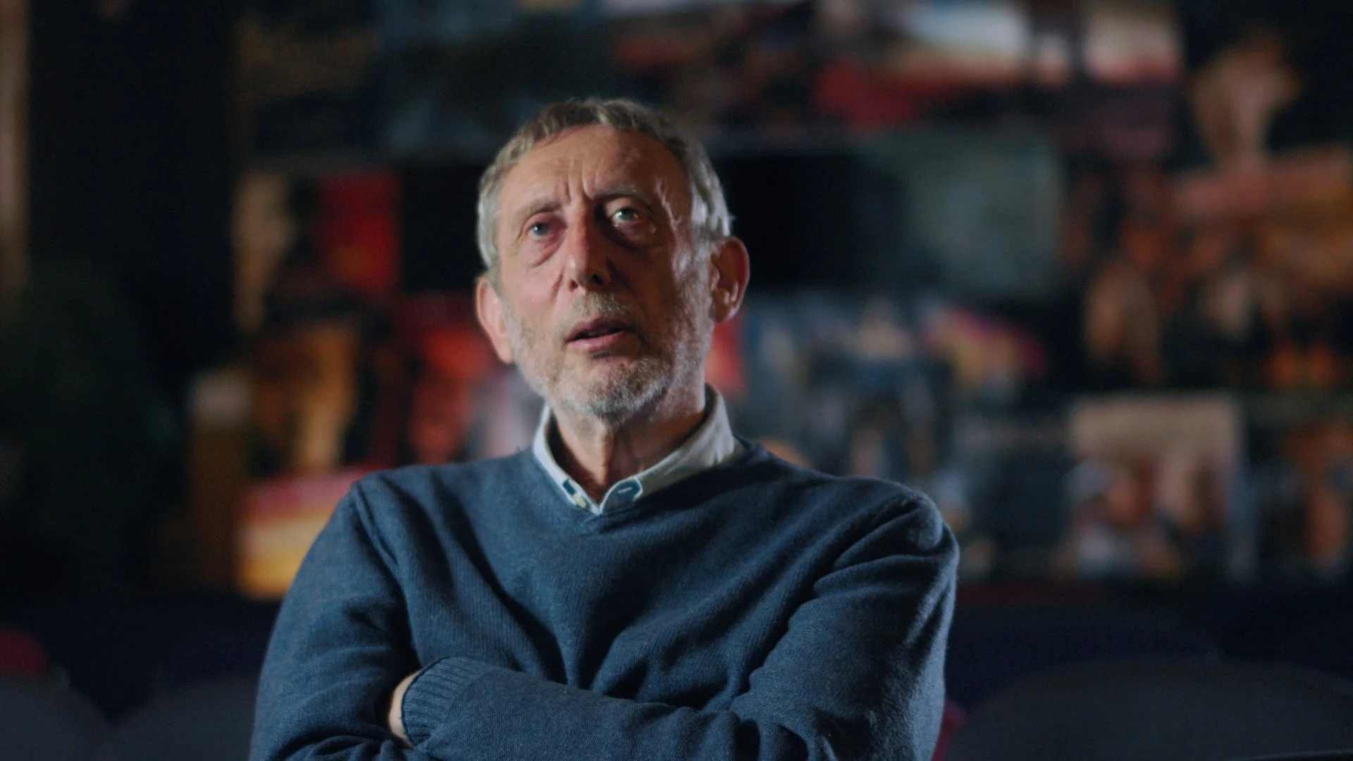 A middle-aged man with gray hair and beard, wearing a blue sweater, sitting with arms crossed in a room with shelves and objects in the background.