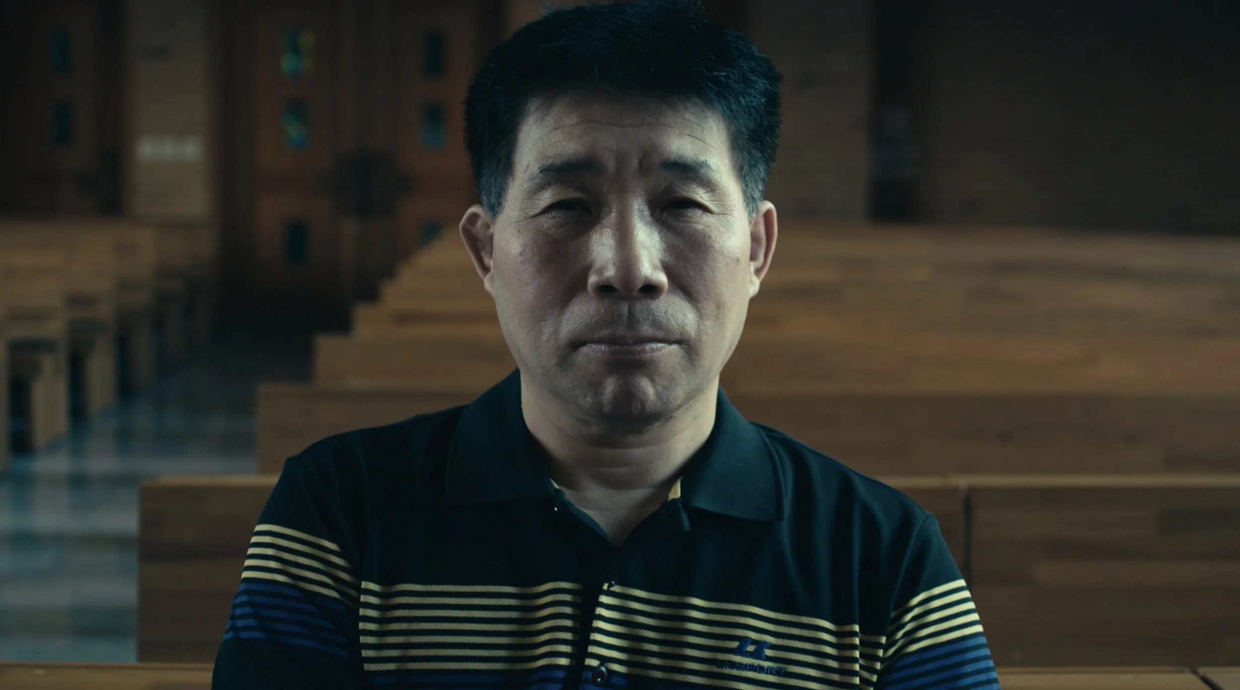 A man with short black hair wearing a navy blue collared shirt with yellow and white stripes on the sleeves sitting on a church pew in a church with wood paneling and pews in the background.