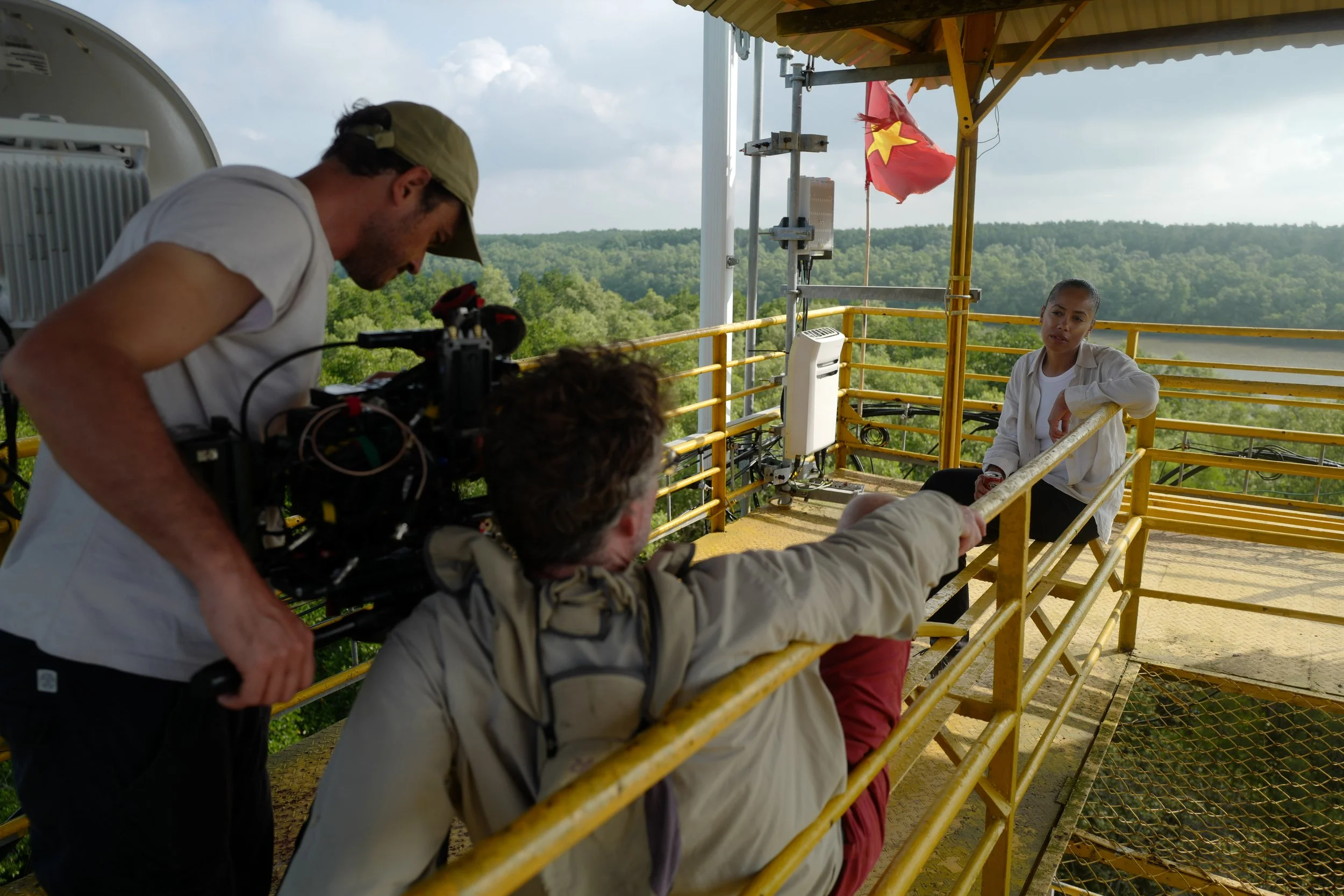 People on an outdoor platform with a woman sitting and speaking, crew members filming her, and a Vietnamese flag in the background.