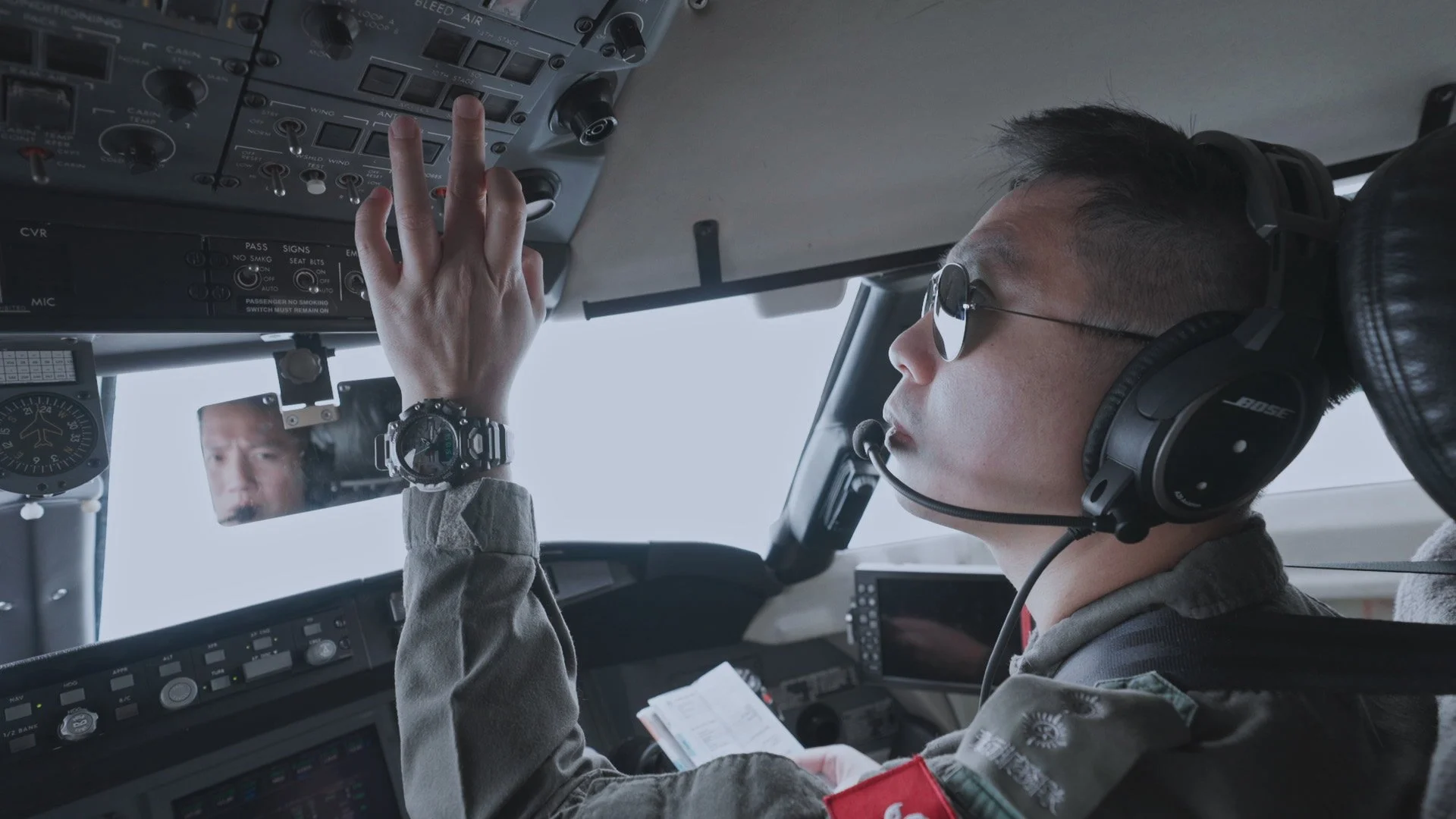 A pilot wearing sunglasses and a headset is sitting in the cockpit, adjusting controls with their hand, with another person reflected in a small mirror.