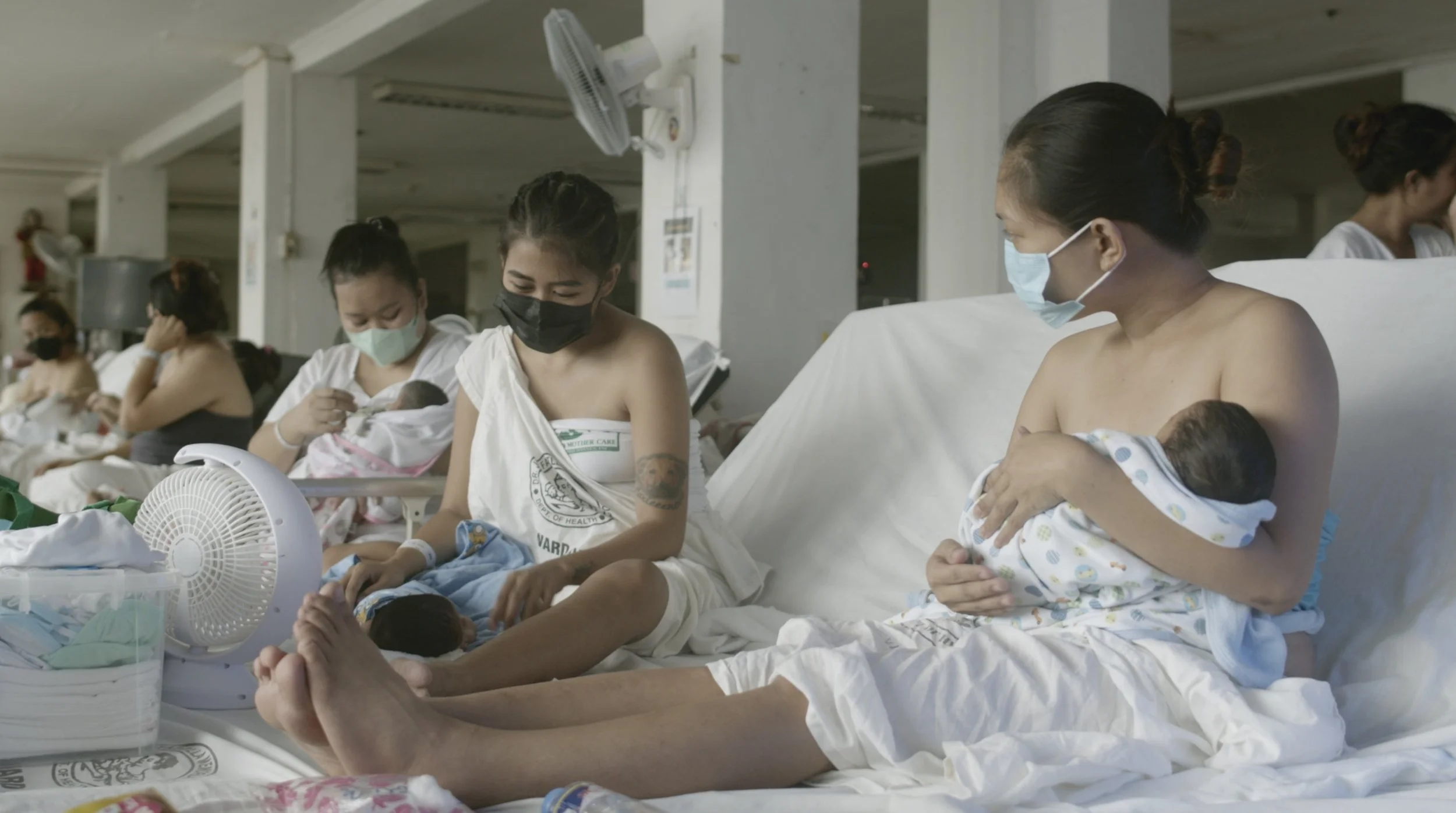 A group of women in a hospital maternity ward, wearing face masks, holding newborn babies, with medical equipment and fans around them.