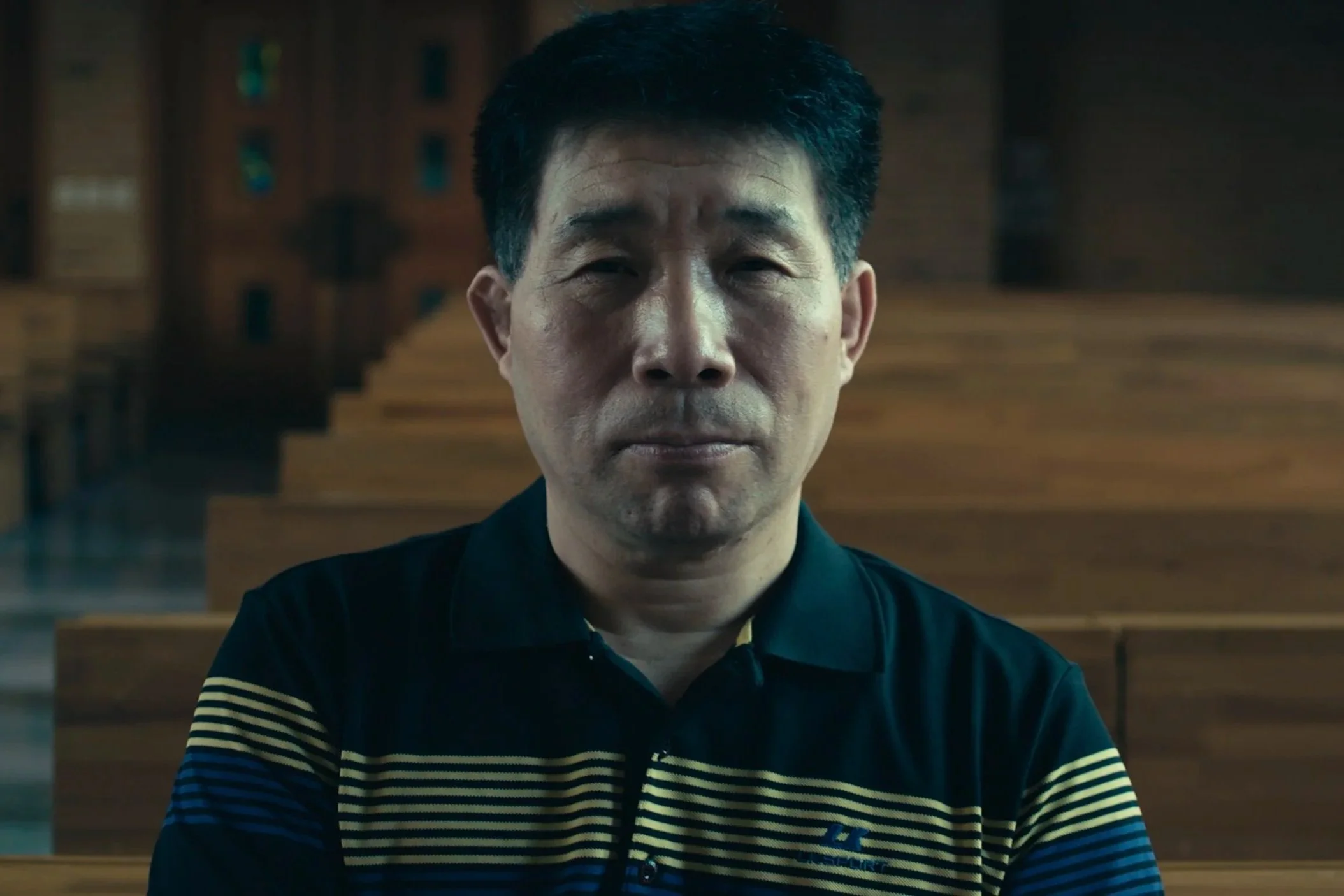 A middle-aged Asian man with short black hair, wearing a black and yellow striped sweater, sitting in a wooden pew inside a church or chapel, with wooden stairs in the background.