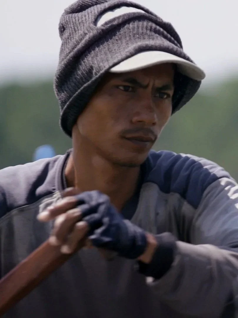 A young man with a serious expression is wearing a gray beanie, a baseball cap, and a gray athletic jacket, and he appears to be involved in a physical activity outdoors.