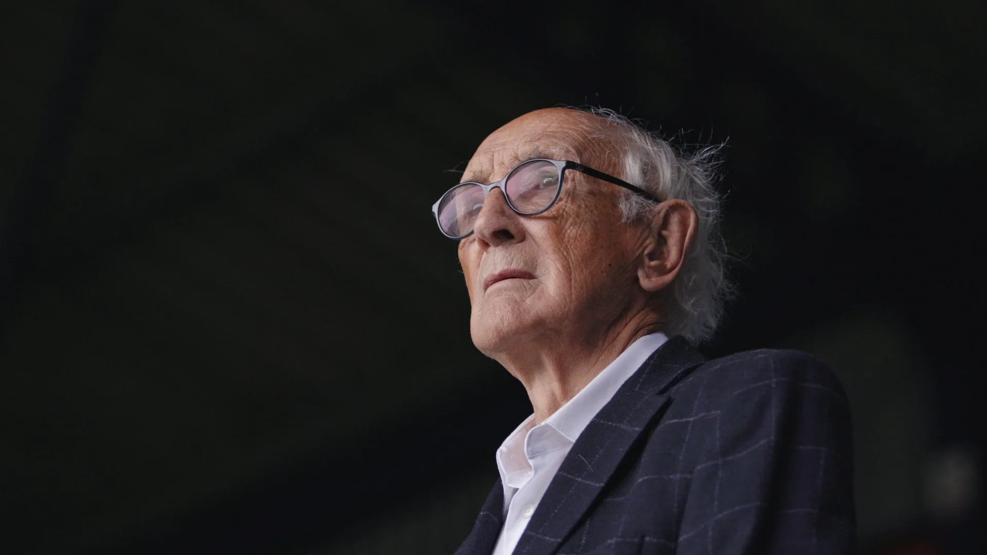 An elderly man wearing glasses and a dark plaid suit looks serious against a dark background, with a slight profile view.