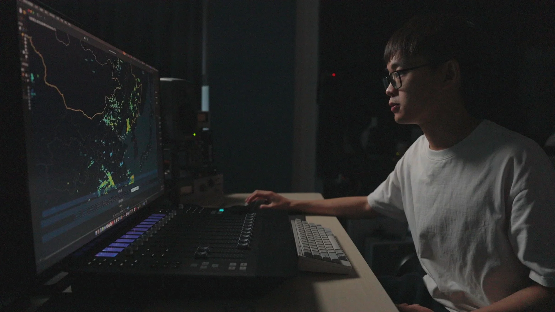 A person wearing glasses and a white shirt working at a music production station with a large computer screen displaying a digital audio workstation and a mixing console.