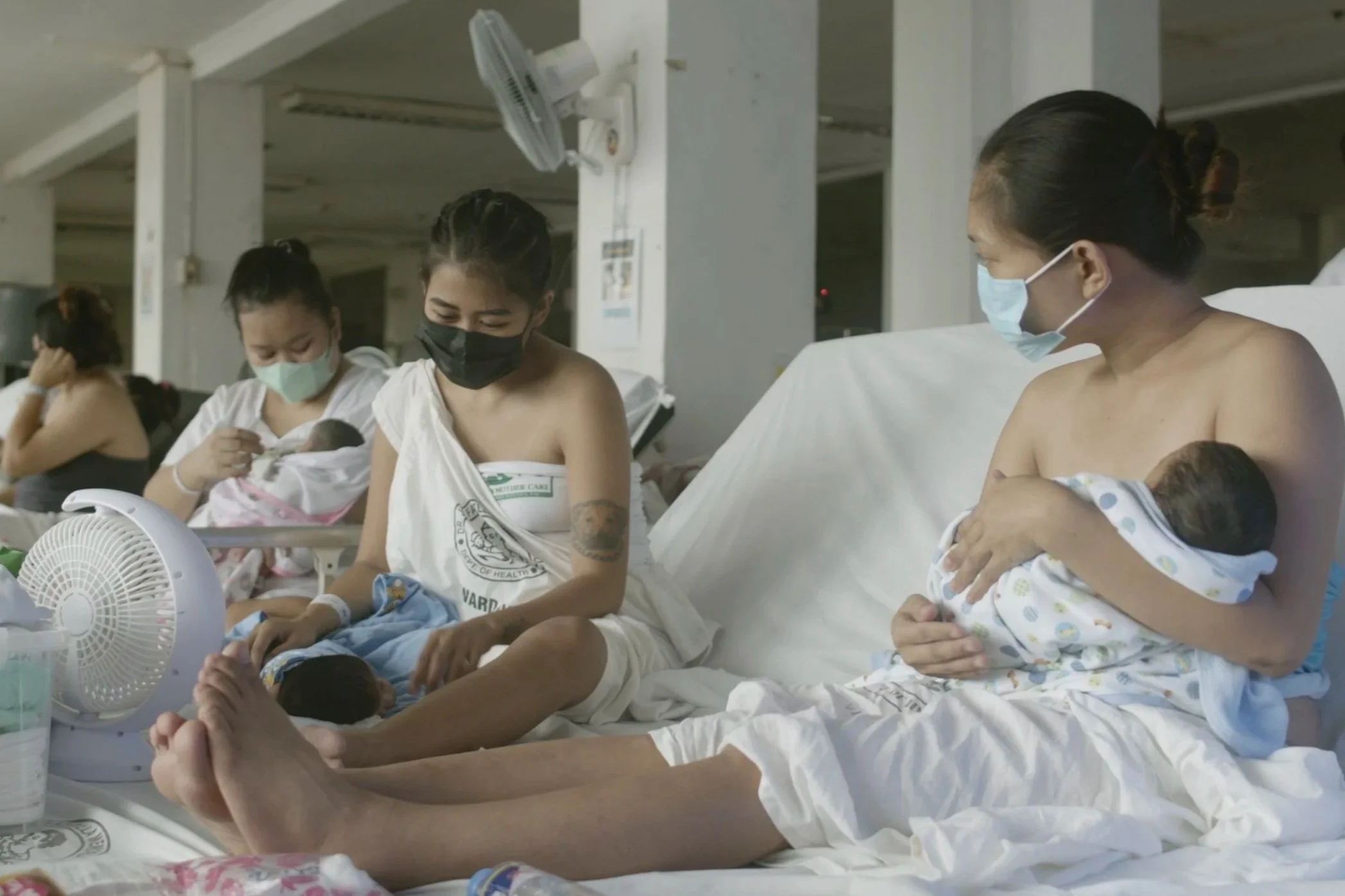 Women wearing masks and hospital gowns, holding newborn babies in a hospital room.