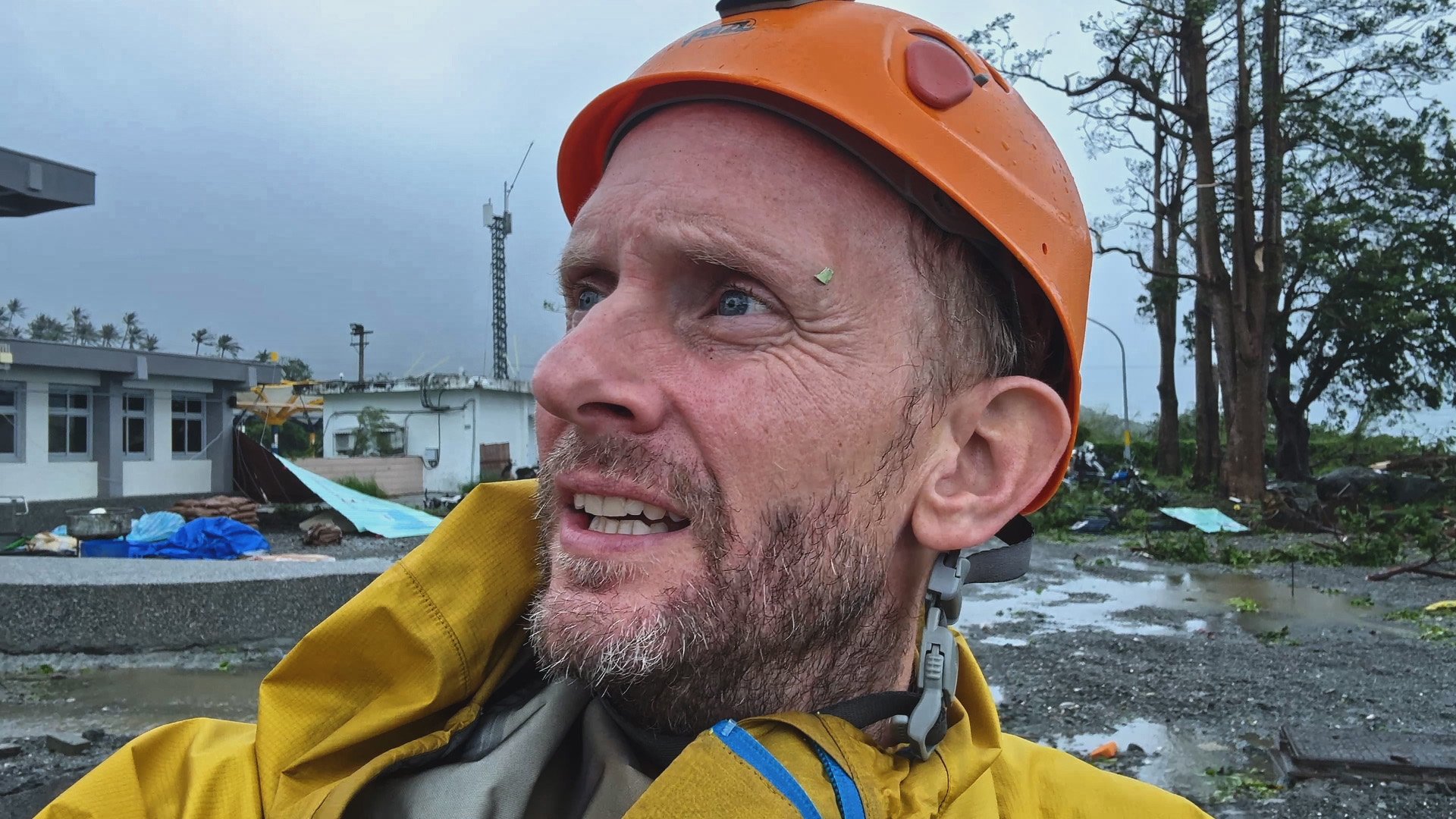 A man wearing an orange safety helmet and yellow raincoat outdoors during a storm, with debris and damaged structures in the background.