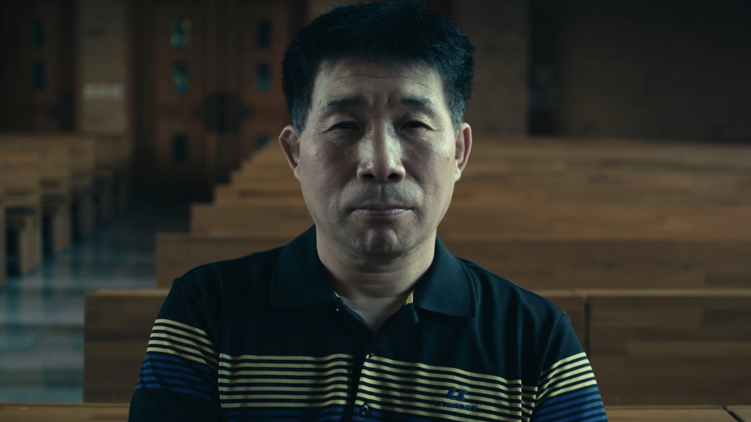 Close-up of a man in a dark shirt with striped yellow and black sleeves, sitting in a large empty church or chapel with wooden pews and stairs in the background.