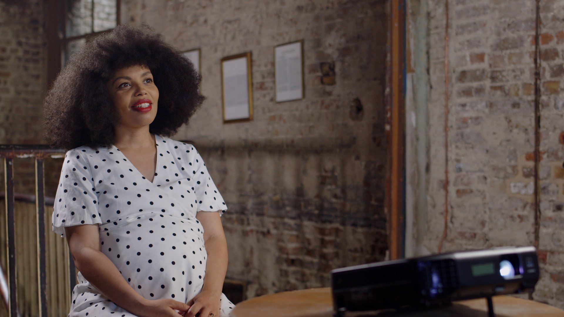 A woman with curly hair, wearing a white dress with black polka dots, smiling and sitting in a room with an exposed brick wall, framed pictures, and a desk in the foreground.