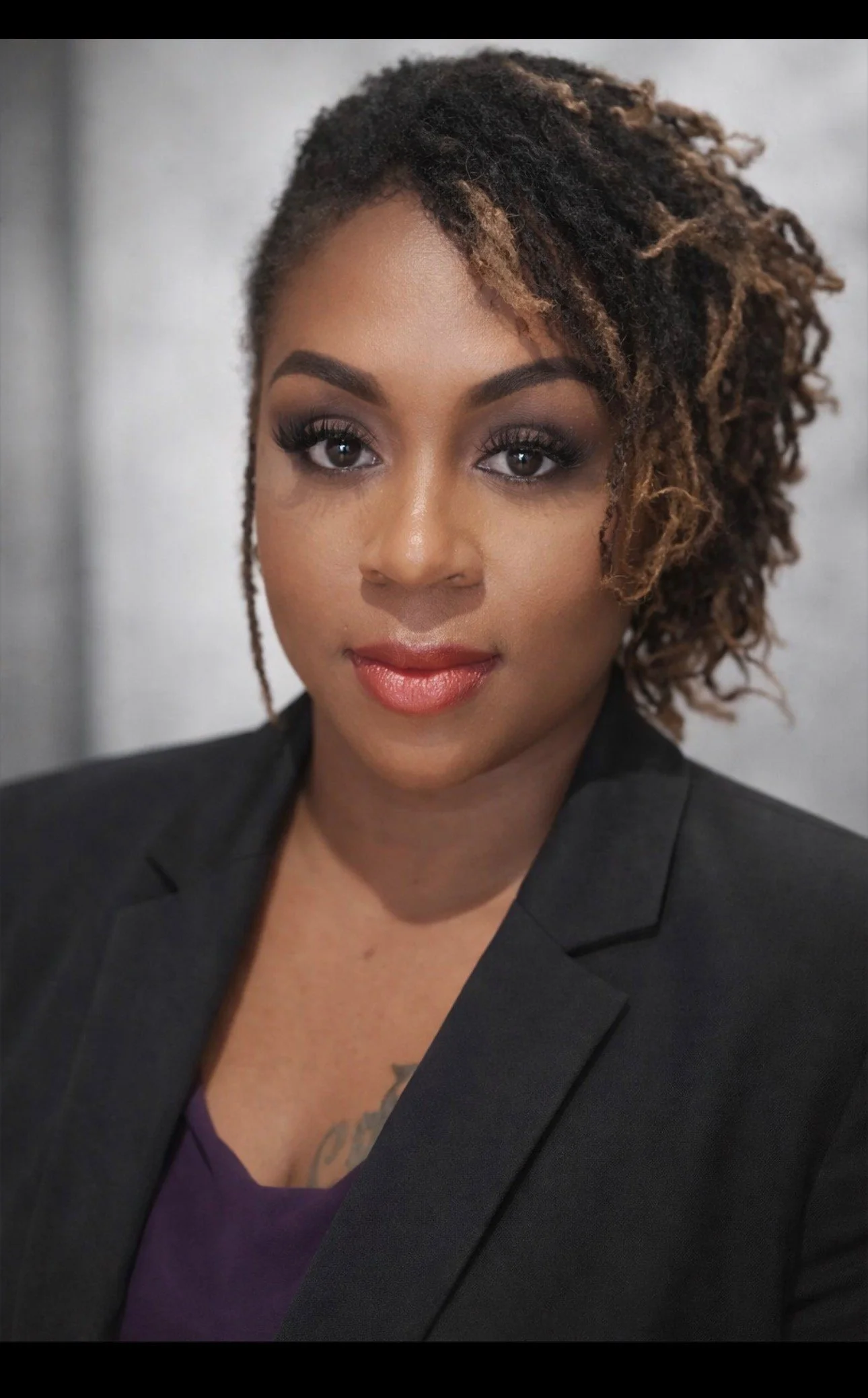 Close-up portrait of a woman with curly brown hair, wearing a dark blazer and a purple top, against a neutral background.
