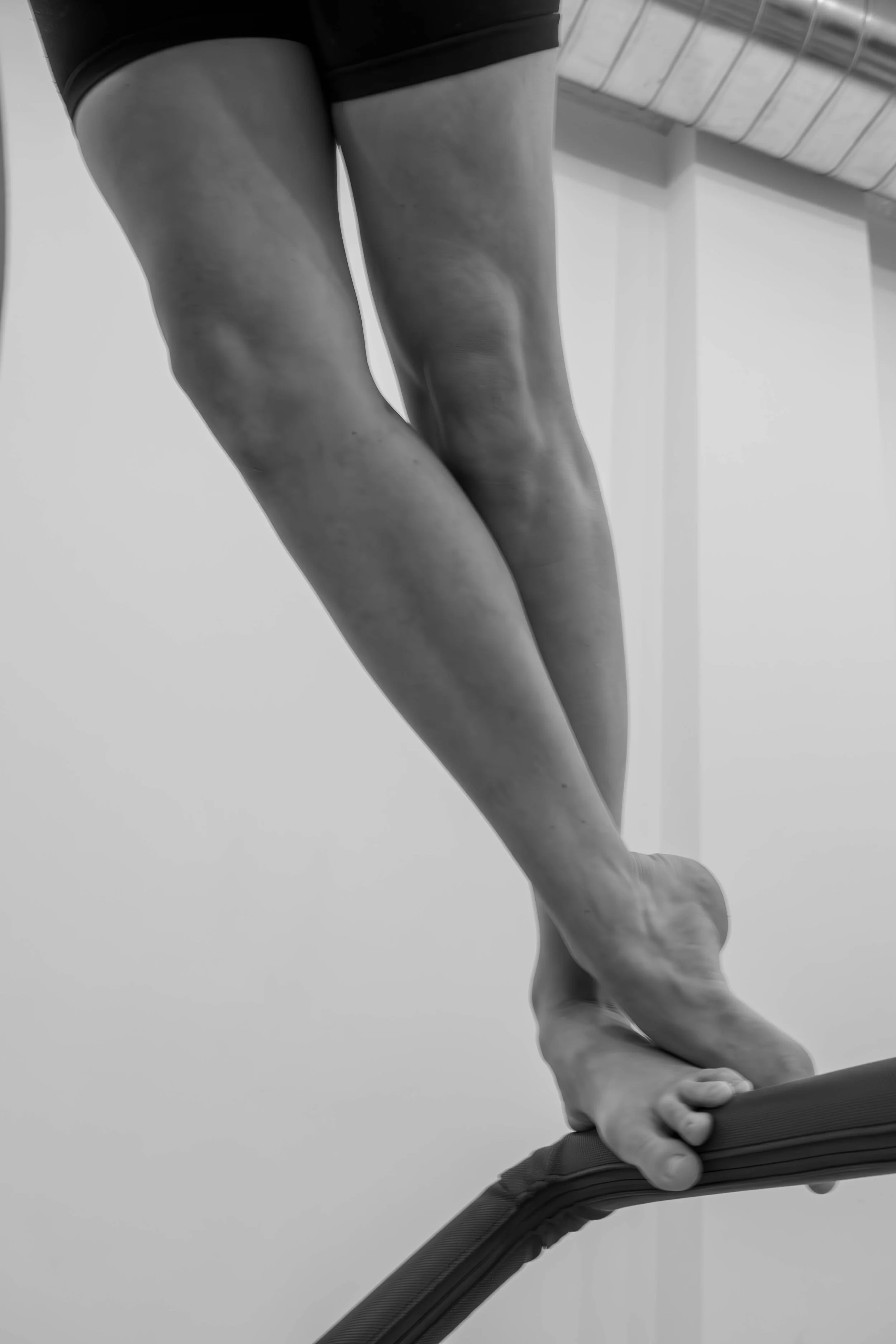 Close-up black and white photo of a person balancing on a ballet barre, wearing shorts, focusing on their legs and hand grip.