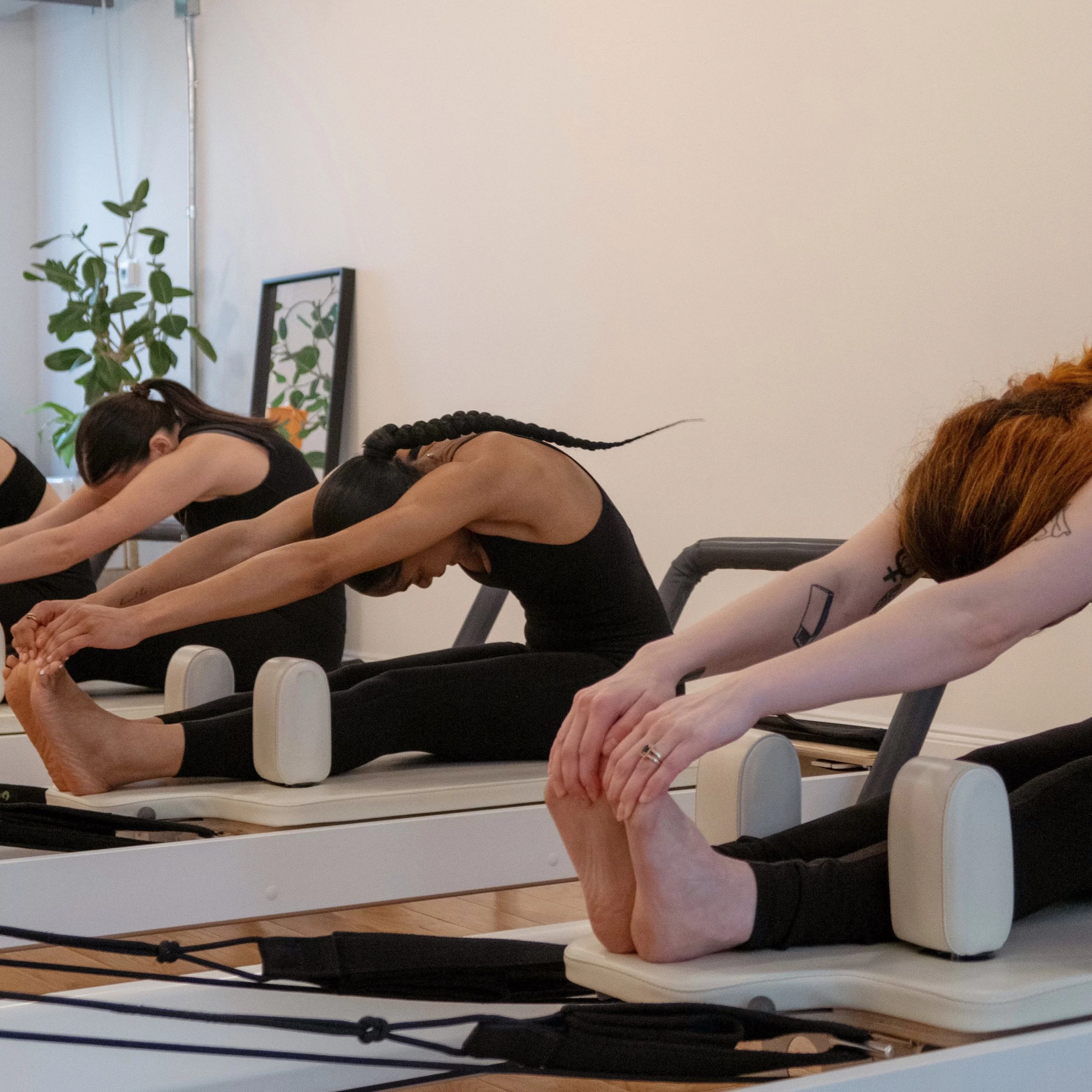 A group of women participating in a Pilates class, stretching on reformer machines in a well-lit studio with a mirror, plants, and wall decorations.