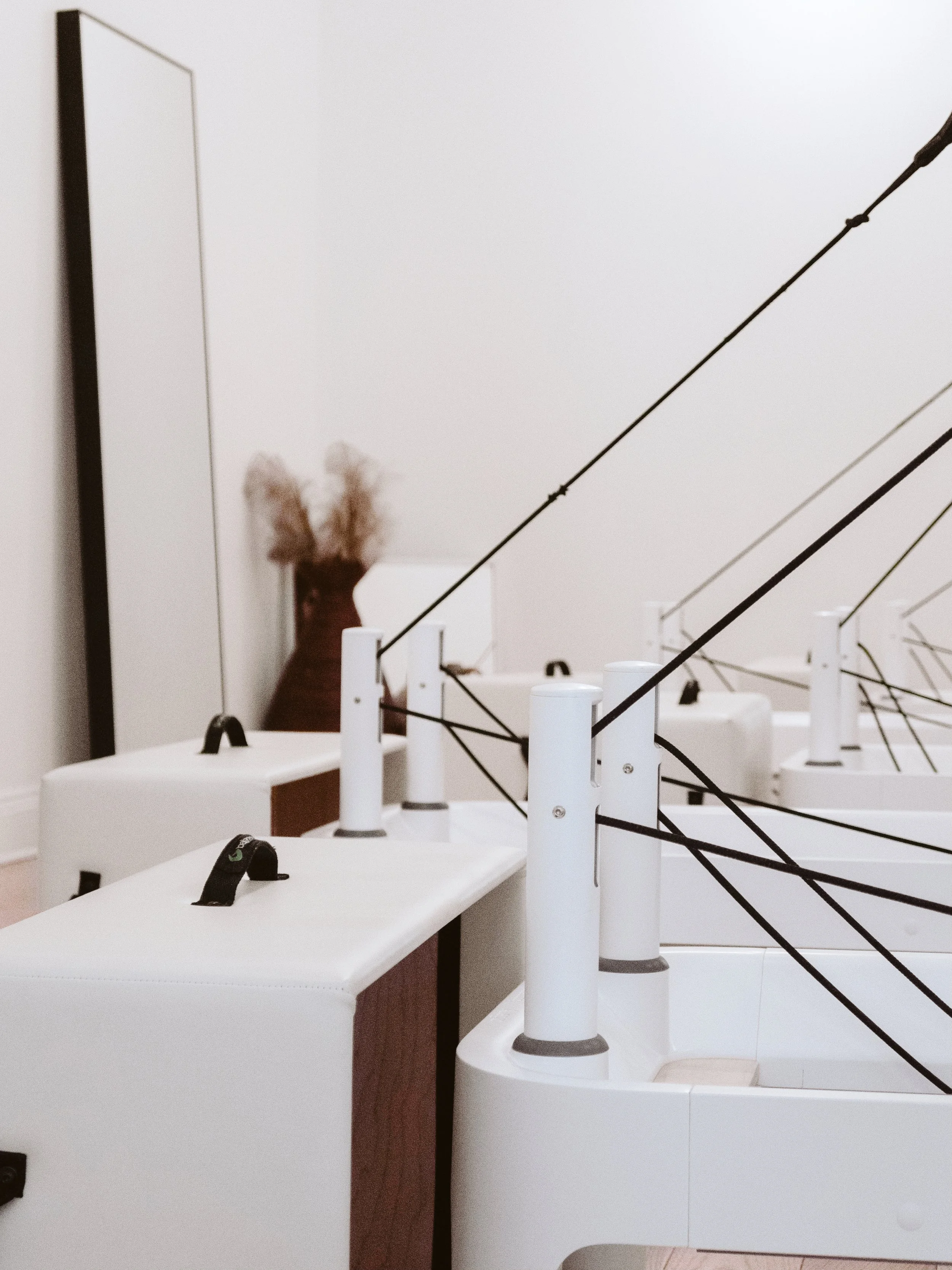 Interior of a Pilates studio with several reformer machines in a minimalist room with a vase of dried pampas grass in the background.