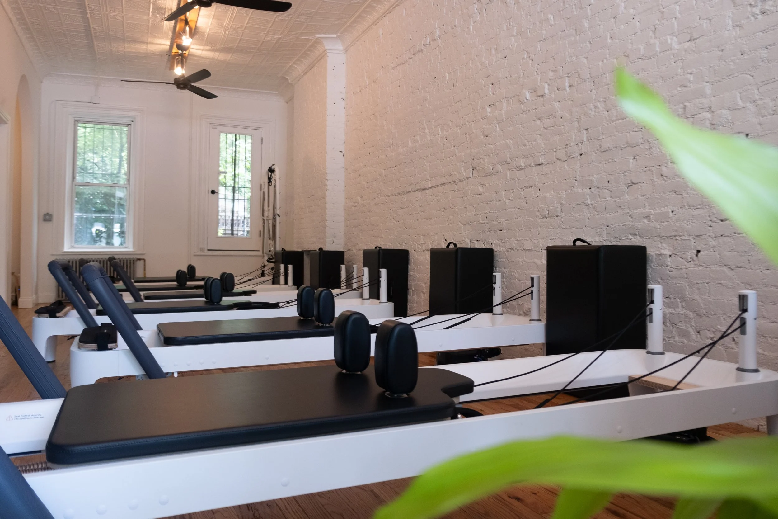 Interior of a Pilates studio with several reformer machines arranged in a row, white brick walls, large windows letting in natural light, ceiling fans, and track lighting.